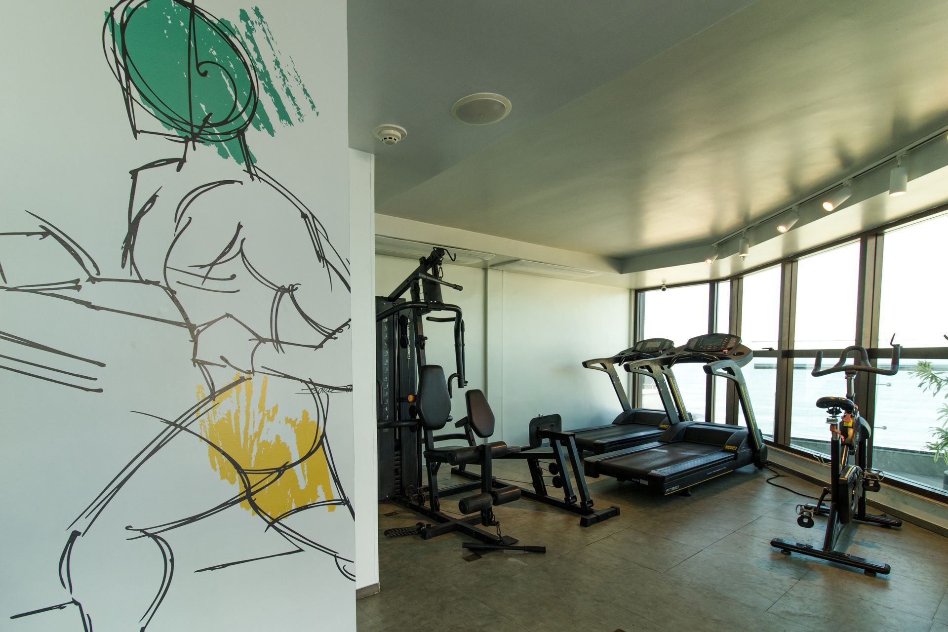 Interior de academia com esteiras, aparelhos de musculação e obras de arte na parede em uma sala bem iluminada.