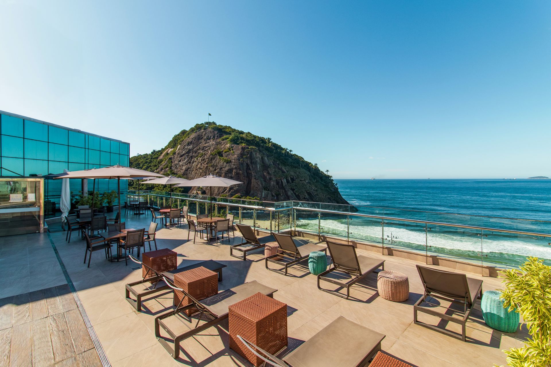 Terraço à beira-mar com espreguiçadeiras, mesas e guarda-sóis junto a um mar turquesa e um promontório rochoso.