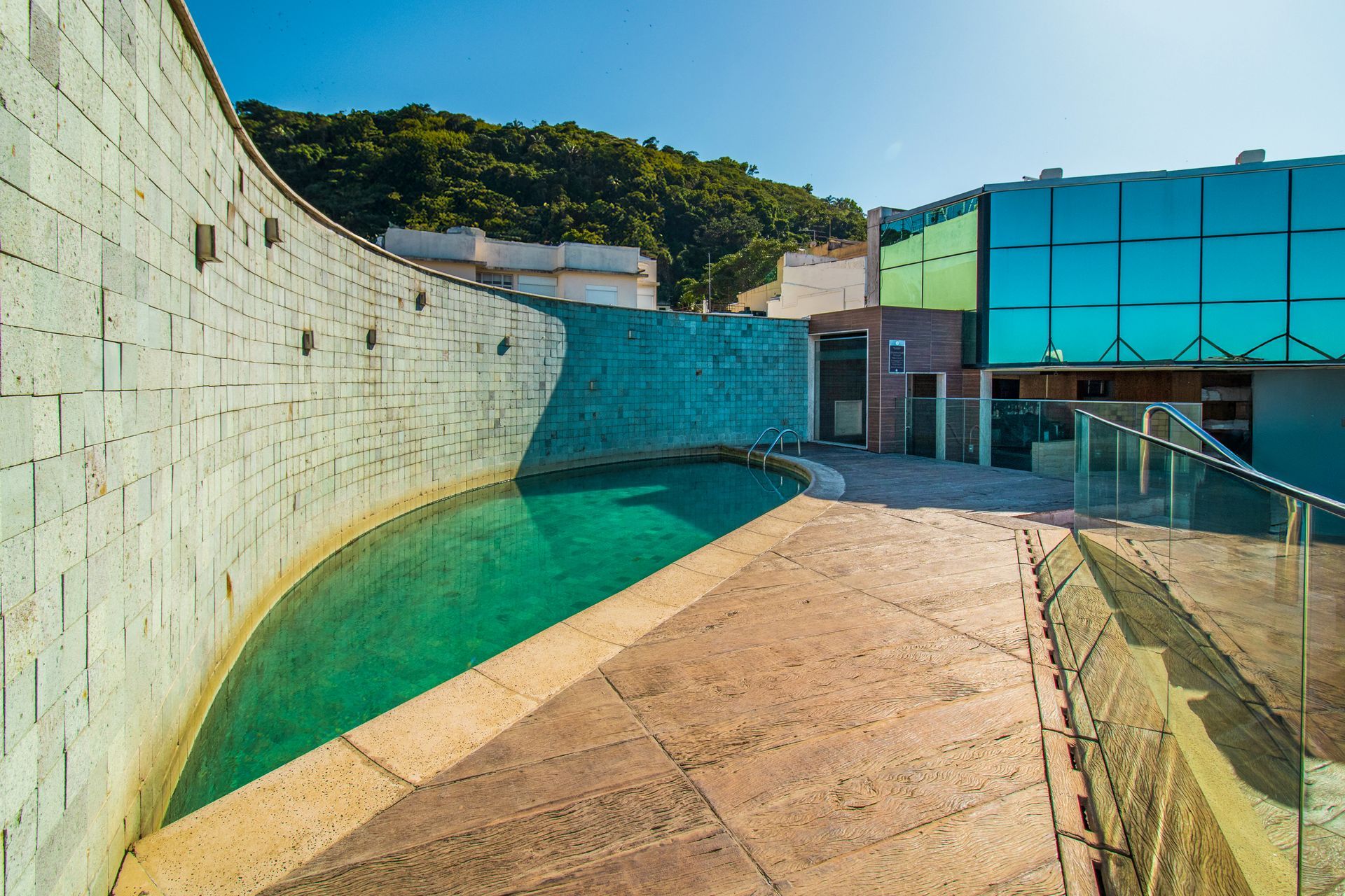Piscina curva turquesa ao lado de um muro de pedra branca e um edifício de vidro sob um céu azul.