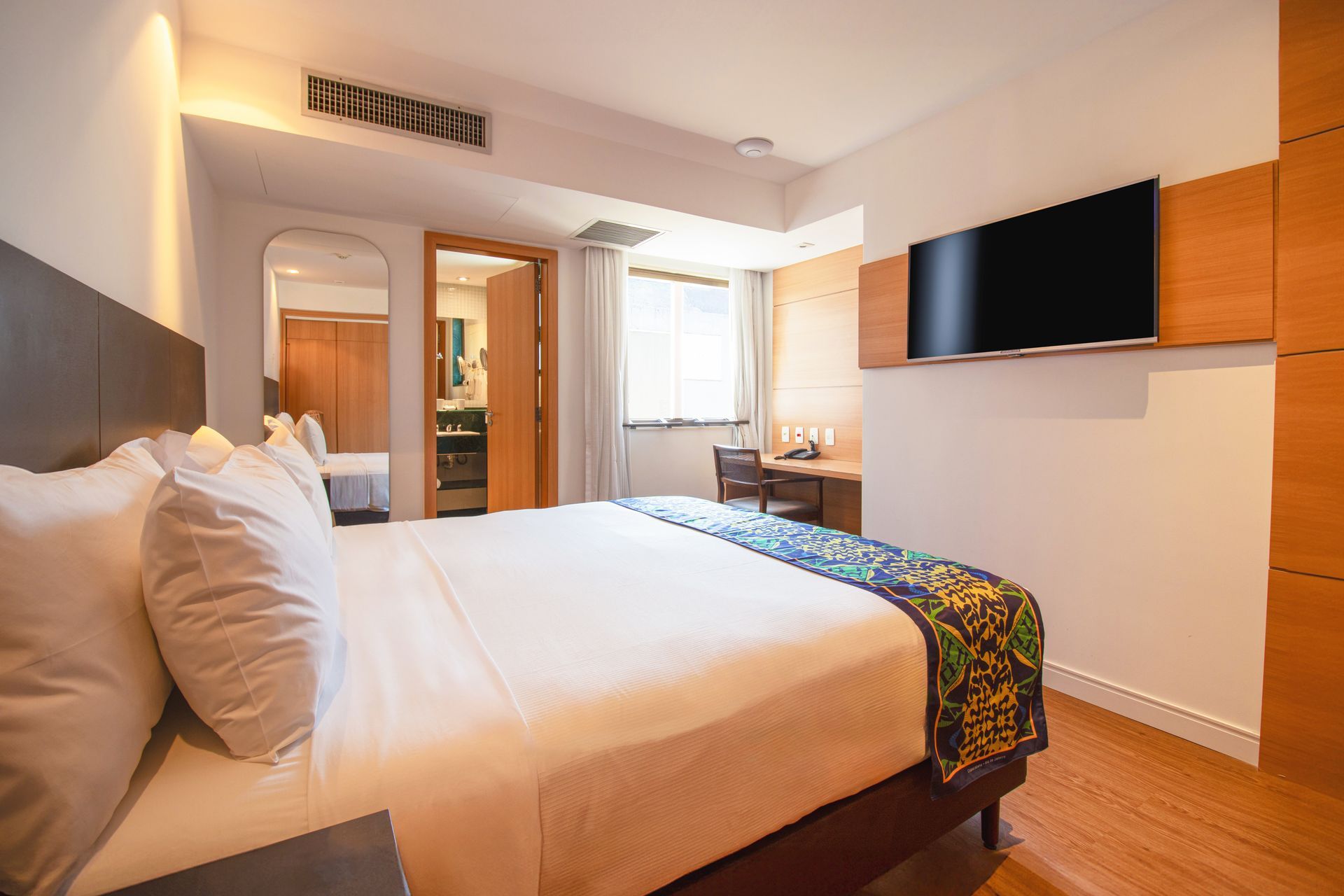 Quarto de hotel moderno com cama, TV de parede e janela com cortinas leves.