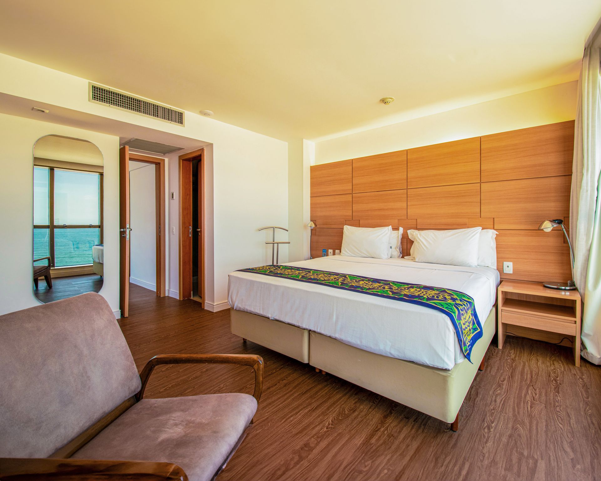 Quarto de hotel luminoso com cama king-size, cabeceira de madeira, poltrona e varanda com vista através de porta aberta.