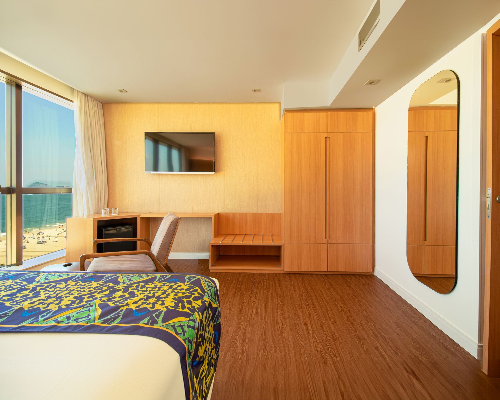 Quarto de hotel moderno com cama, TV de parede, móveis de madeira e uma grande janela com vista para o mar.