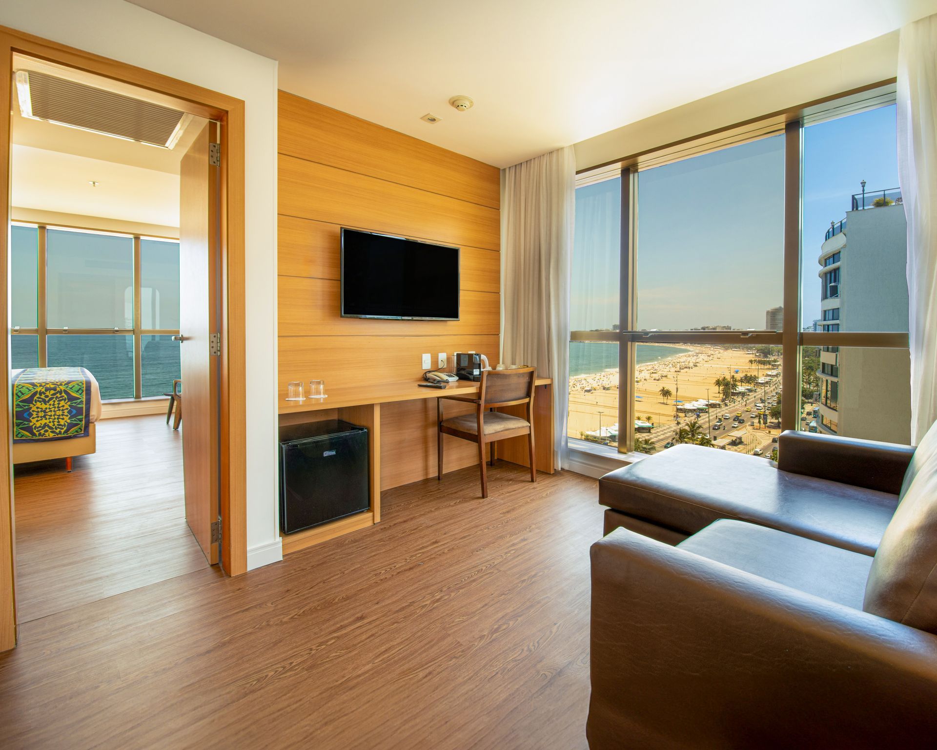 Quarto de hotel luminoso com vista para o mar, TV de parede, escrivaninha e poltrona marrom junto a grandes janelas.