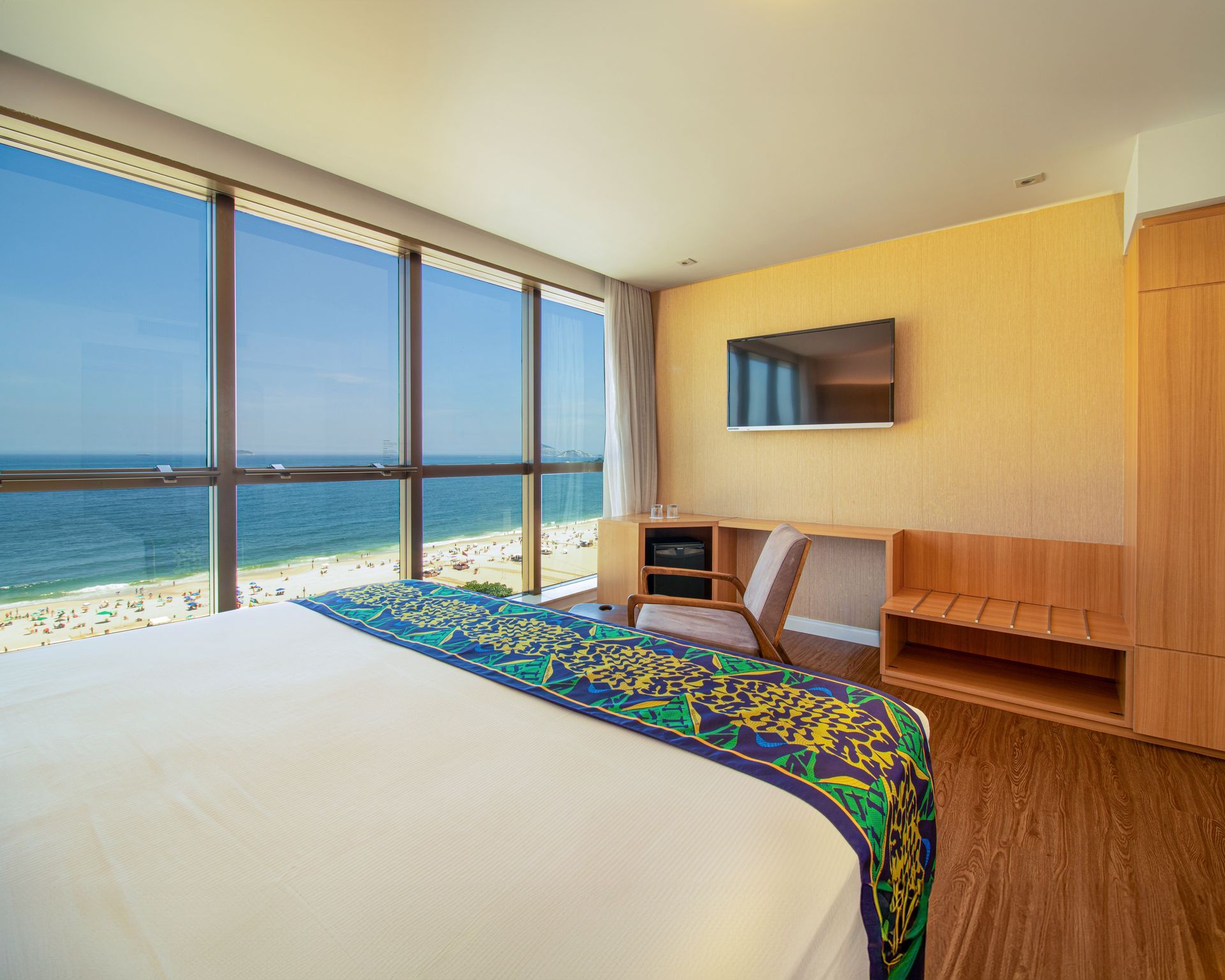 Quarto moderno à beira-mar com janelas amplas, vista para o oceano, cama, TV de parede e painéis de madeira em tons quentes.