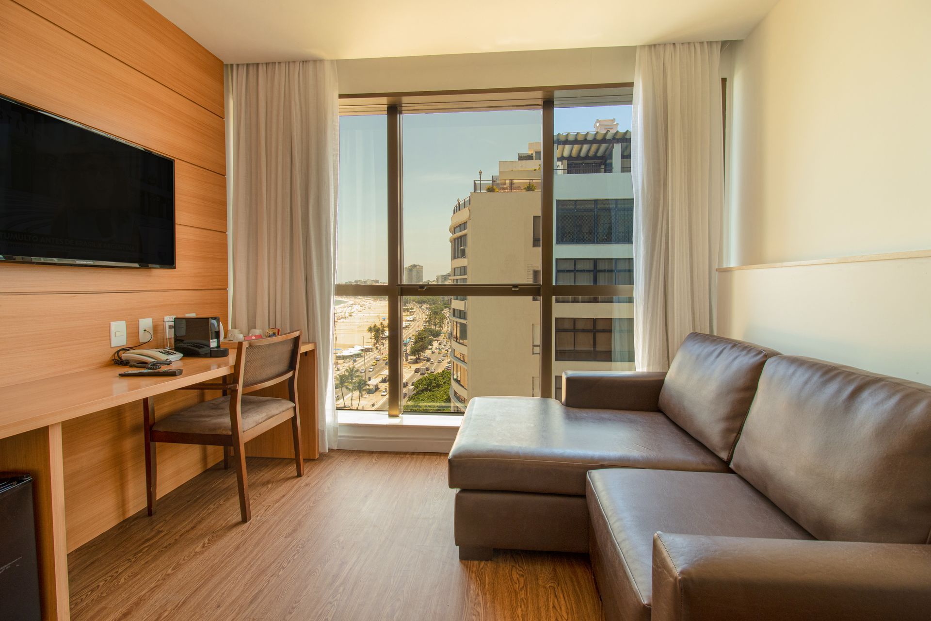 Quarto de hotel moderno com escrivaninha, sofá e janela ampla com vista para os prédios da cidade.