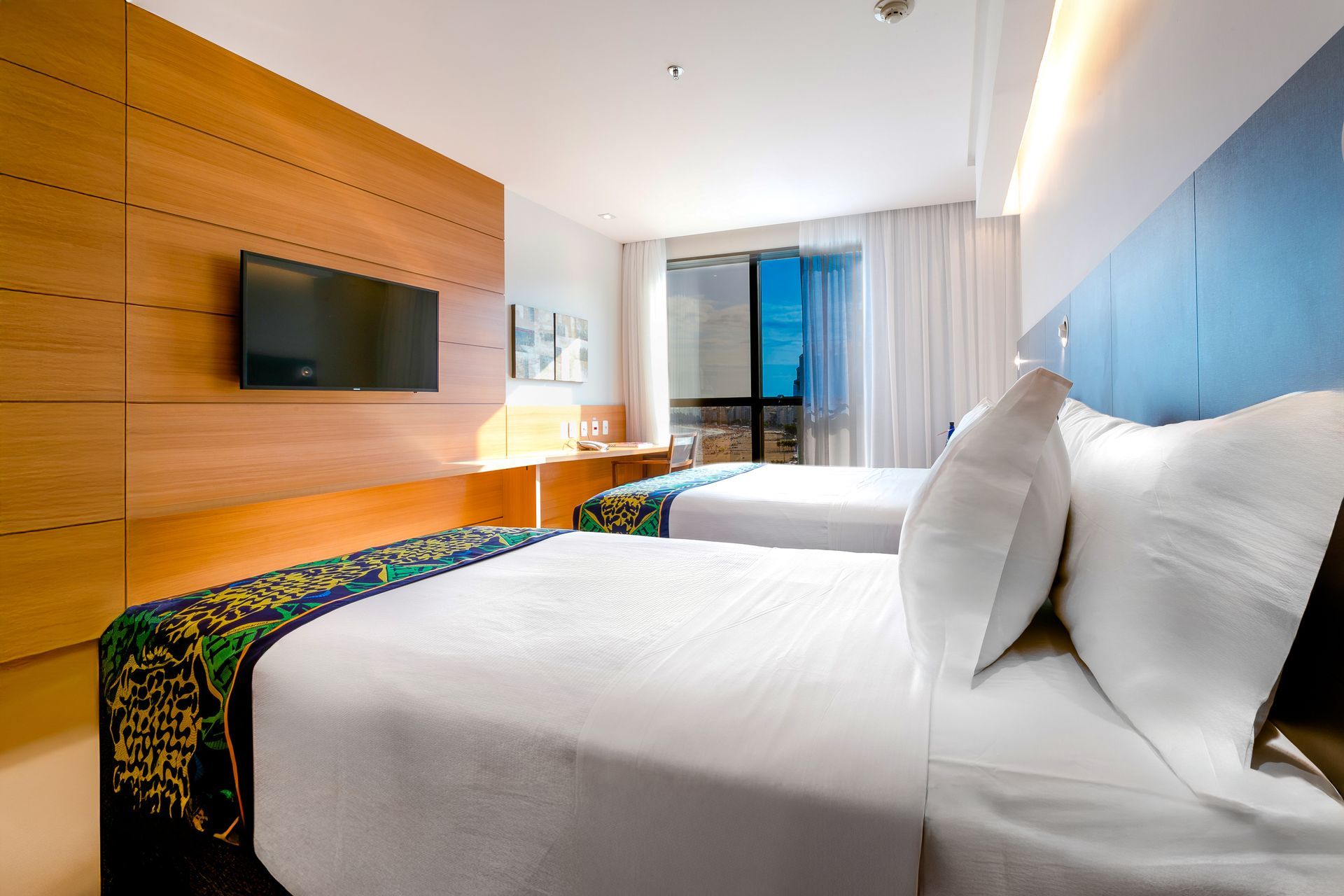 Quarto de hotel moderno com duas camas brancas, parede com detalhes em azul, TV de parede e portas para varanda.