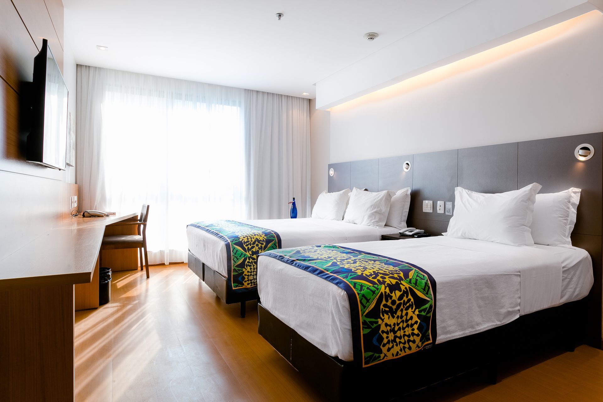 Quarto de hotel moderno com duas camas, janela ampla, piso de madeira e TV de parede.