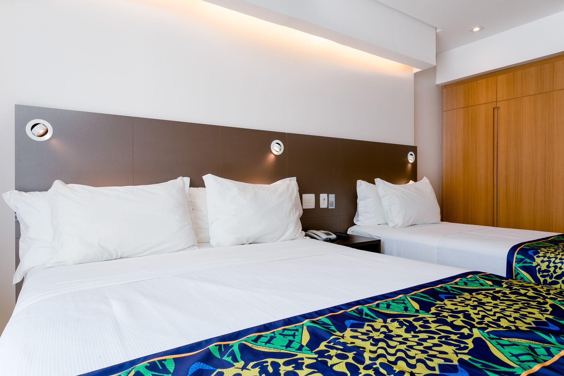 Quarto de hotel moderno com três camas brancas e uma peseira azul estampada.