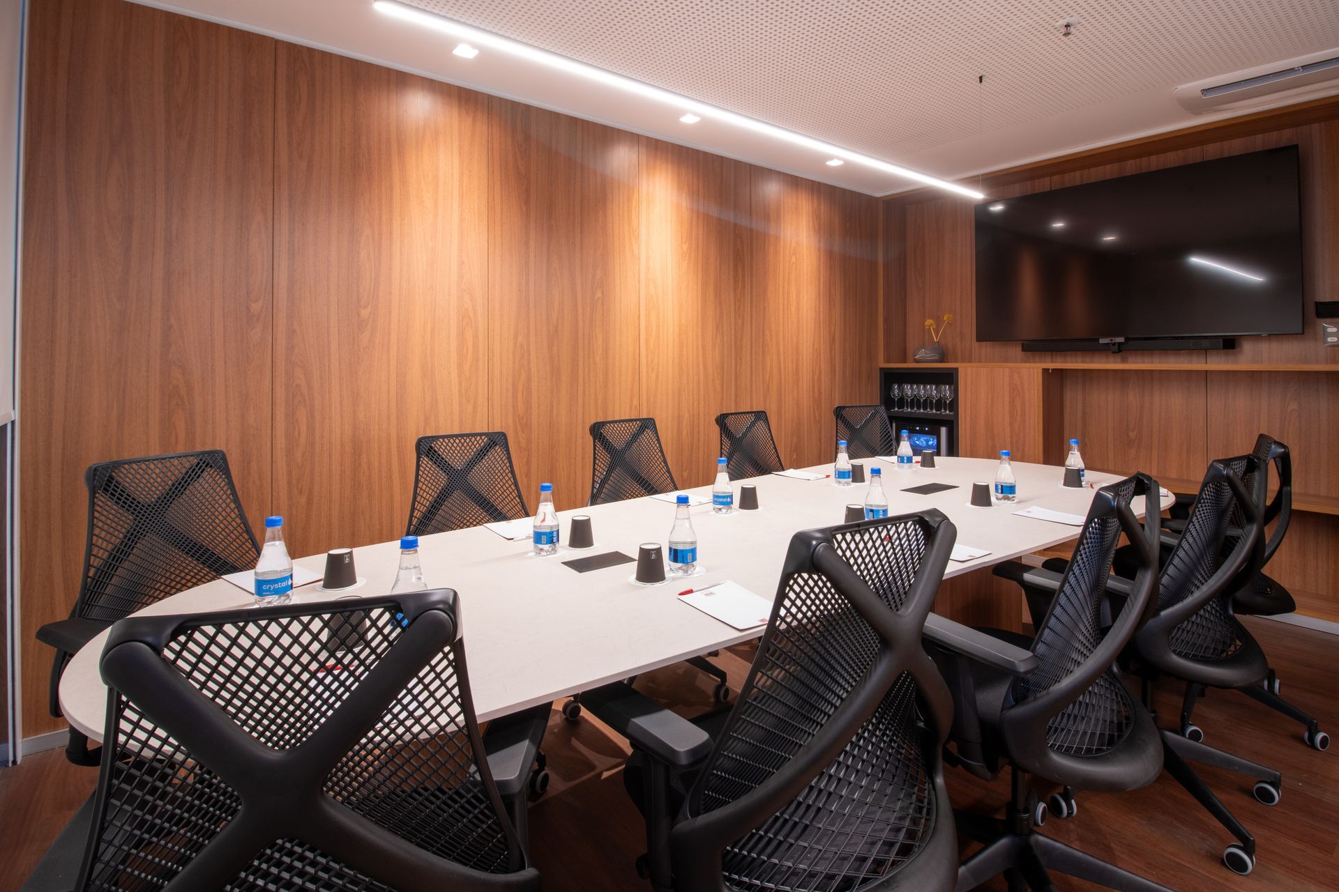 Sala de conferências moderna com mesa comprida branca, cadeiras pretas, paredes revestidas de madeira e uma TV fixada na parede.