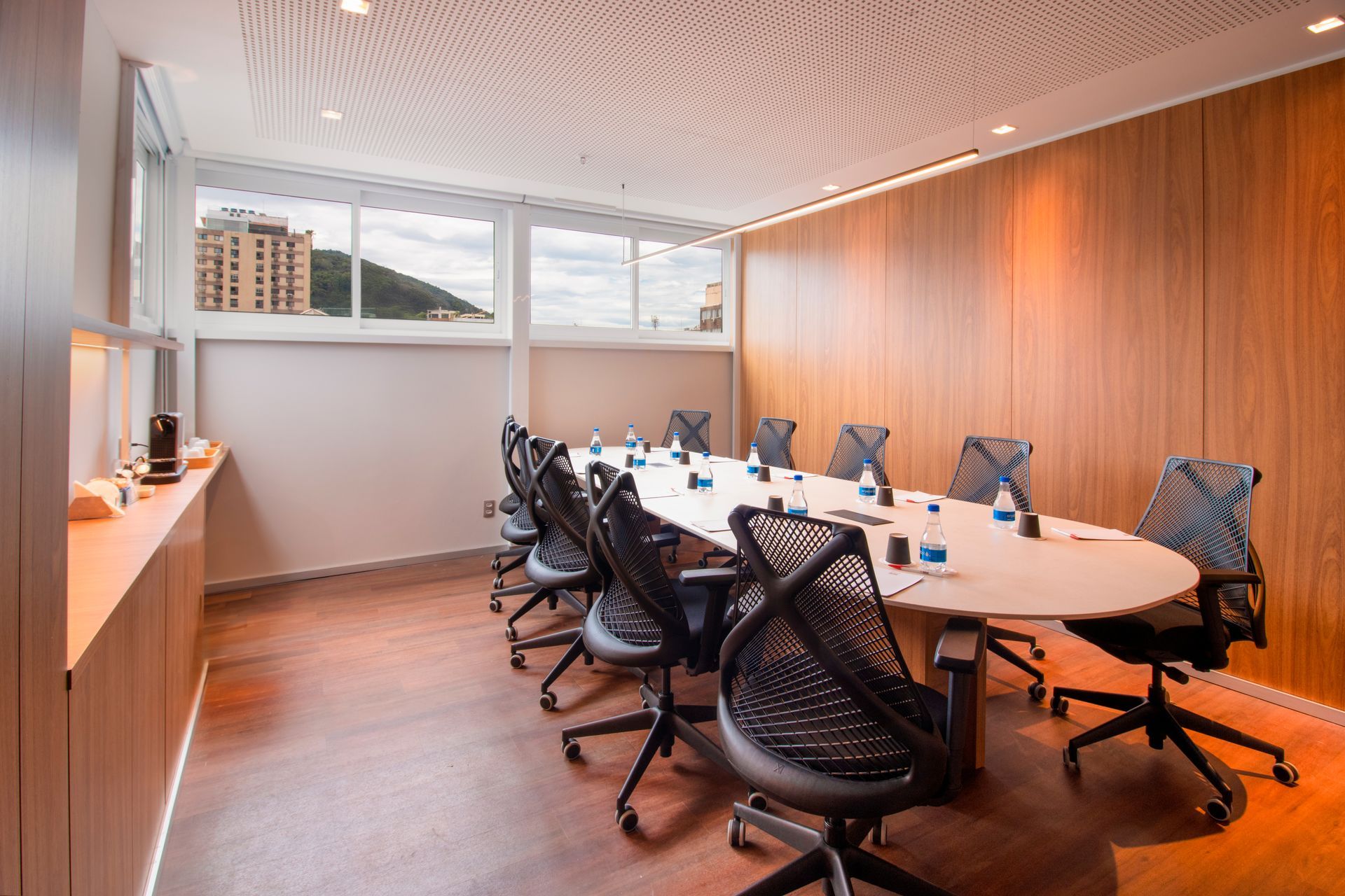 Sala de conferências moderna com mesa oval, cadeiras de escritório pretas, paredes de madeira e grandes janelas.