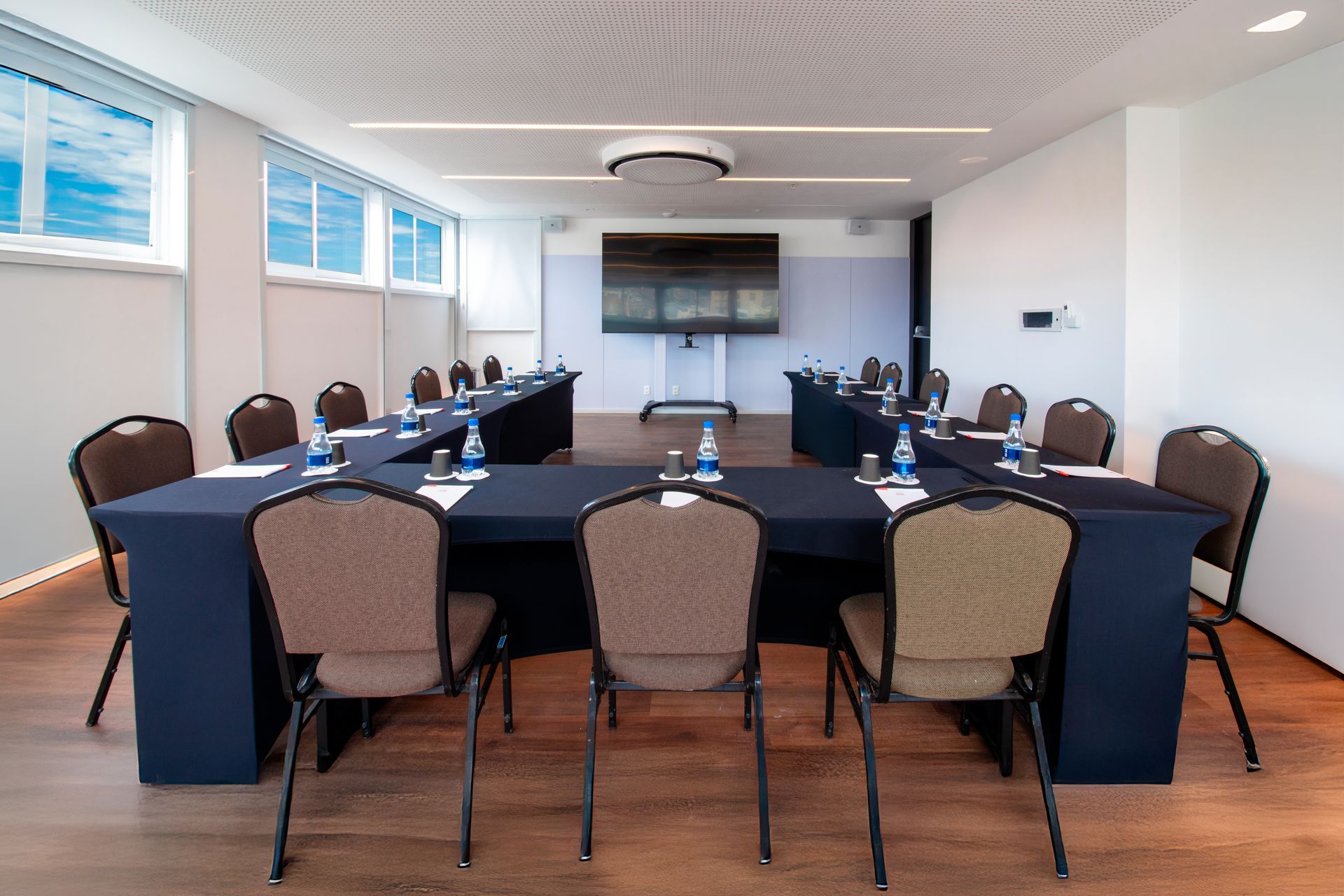 Sala de conferências vazia com mesas em formato de U, cadeiras pretas, TV fixada na parede e janelas grandes.