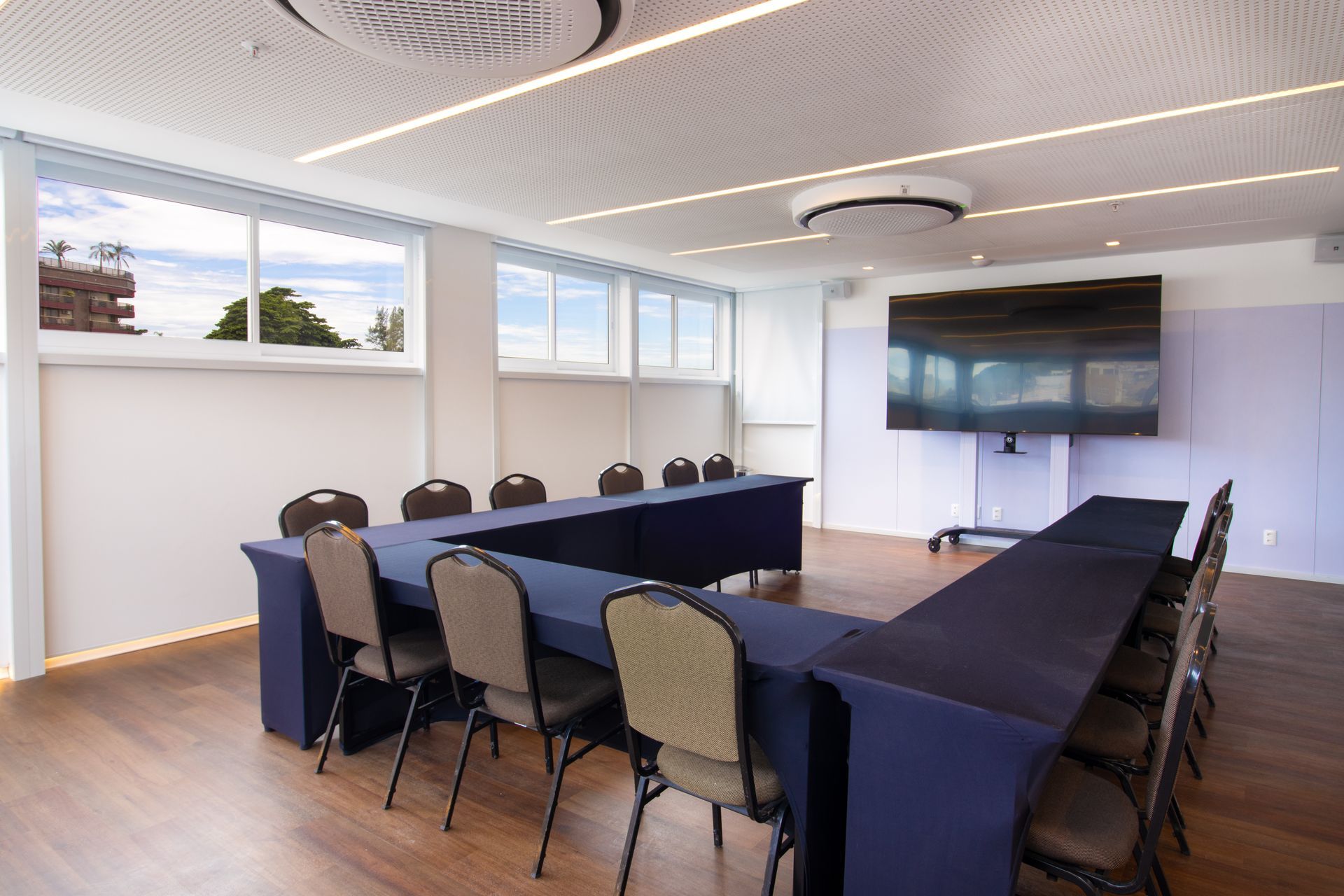 Sala de conferências moderna com mesa em formato de U, cadeiras, TV fixada na parede e janelas amplas.