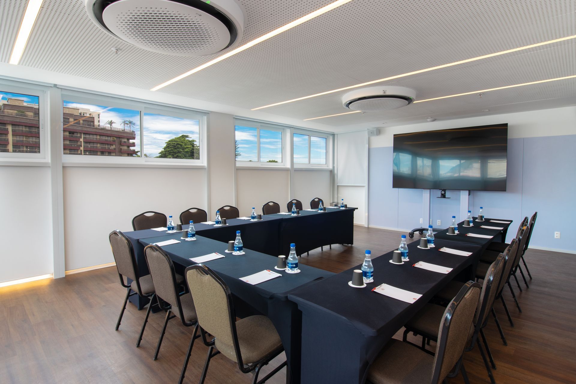 Sala de conferências moderna com uma longa mesa preta, cadeiras, TV fixada na parede e janelas amplas.
