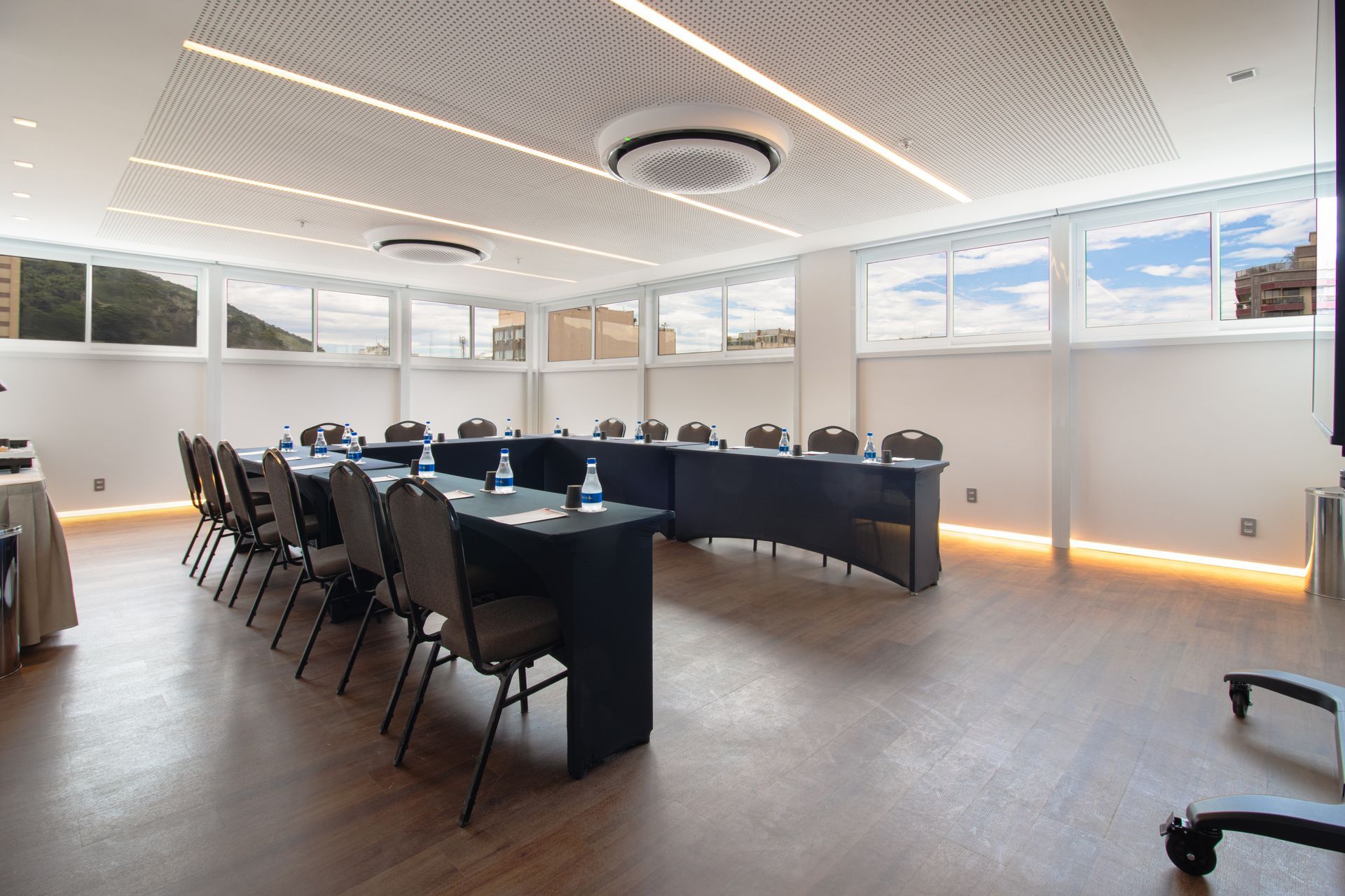 Sala de conferências com mesas pretas em formato de U, cadeiras vazias e janelas amplas com piso de madeira.