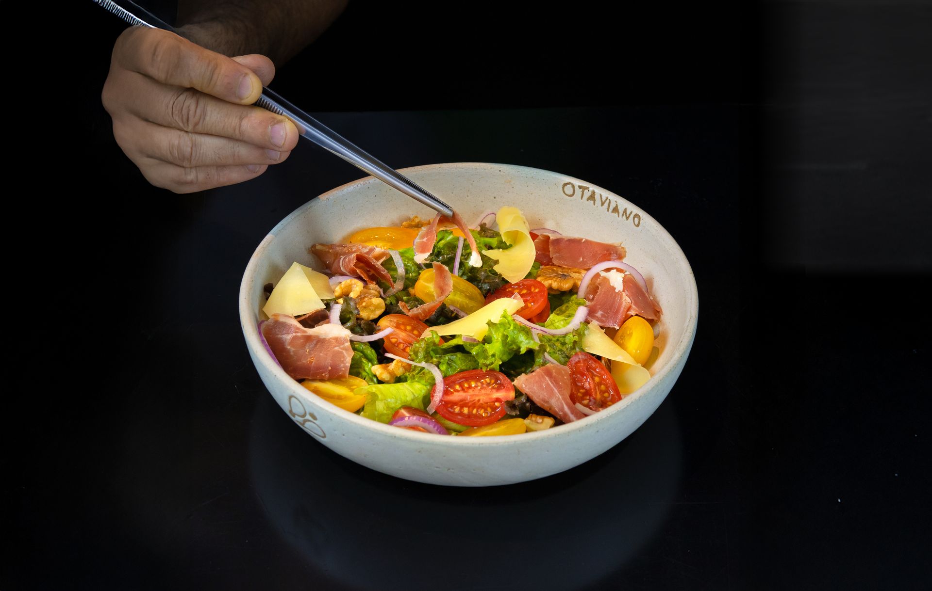 Mão segurando um garfo sobre uma tigela de salada colorida com tomates, pepinos e frutas em uma mesa escura.