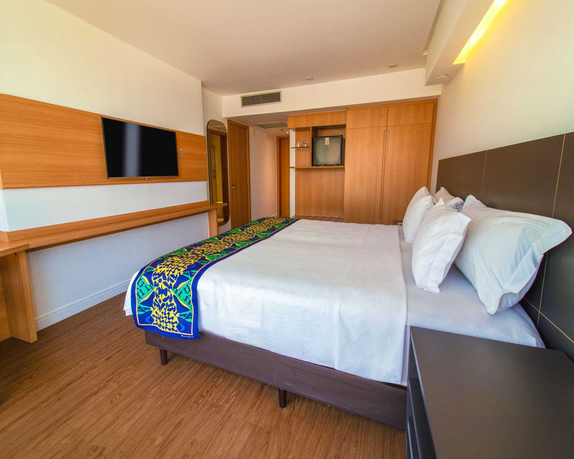 Quarto de hotel moderno com cama, escrivaninha, TV de parede e armário de madeira em tons quentes.
