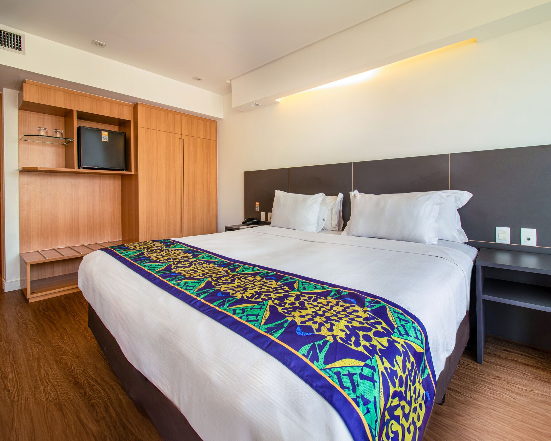 Quarto de hotel moderno com cama grande, tapete azul estampado, móvel de madeira para TV e paredes brancas.