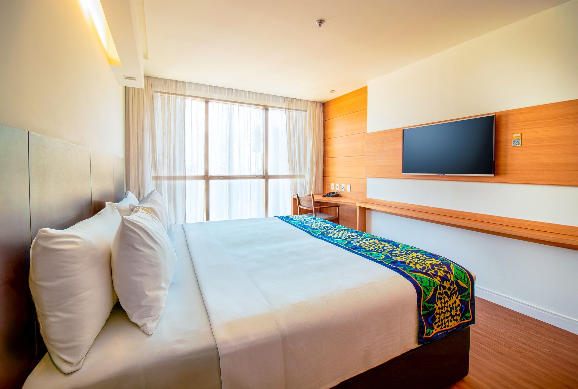 Quarto de hotel moderno com cama king-size, TV de parede, detalhes em madeira e cortinas amplas nas janelas.