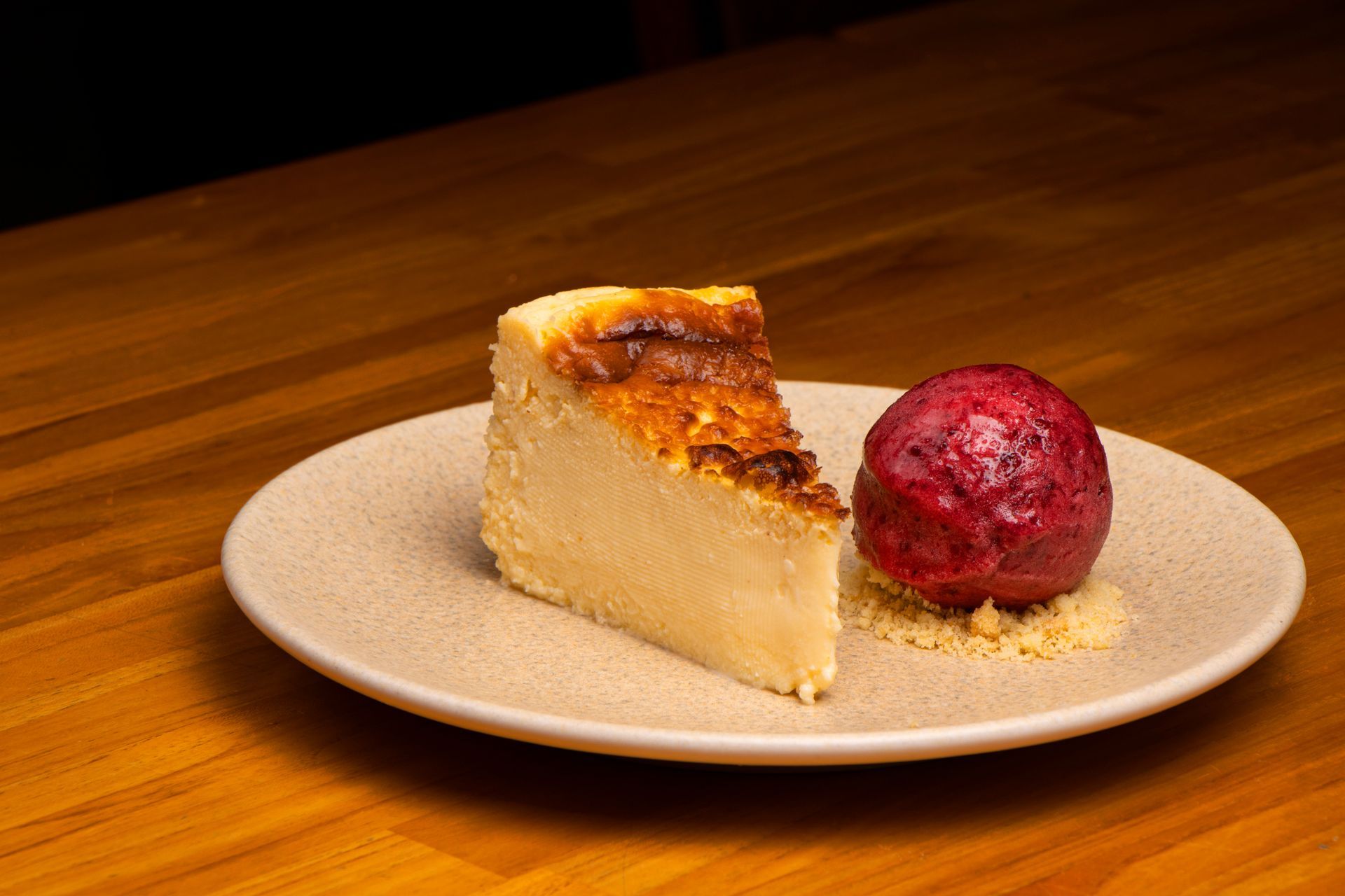 Fatia de cheesecake com sorvete de frutas vermelhas em um prato bege sobre uma mesa de madeira.