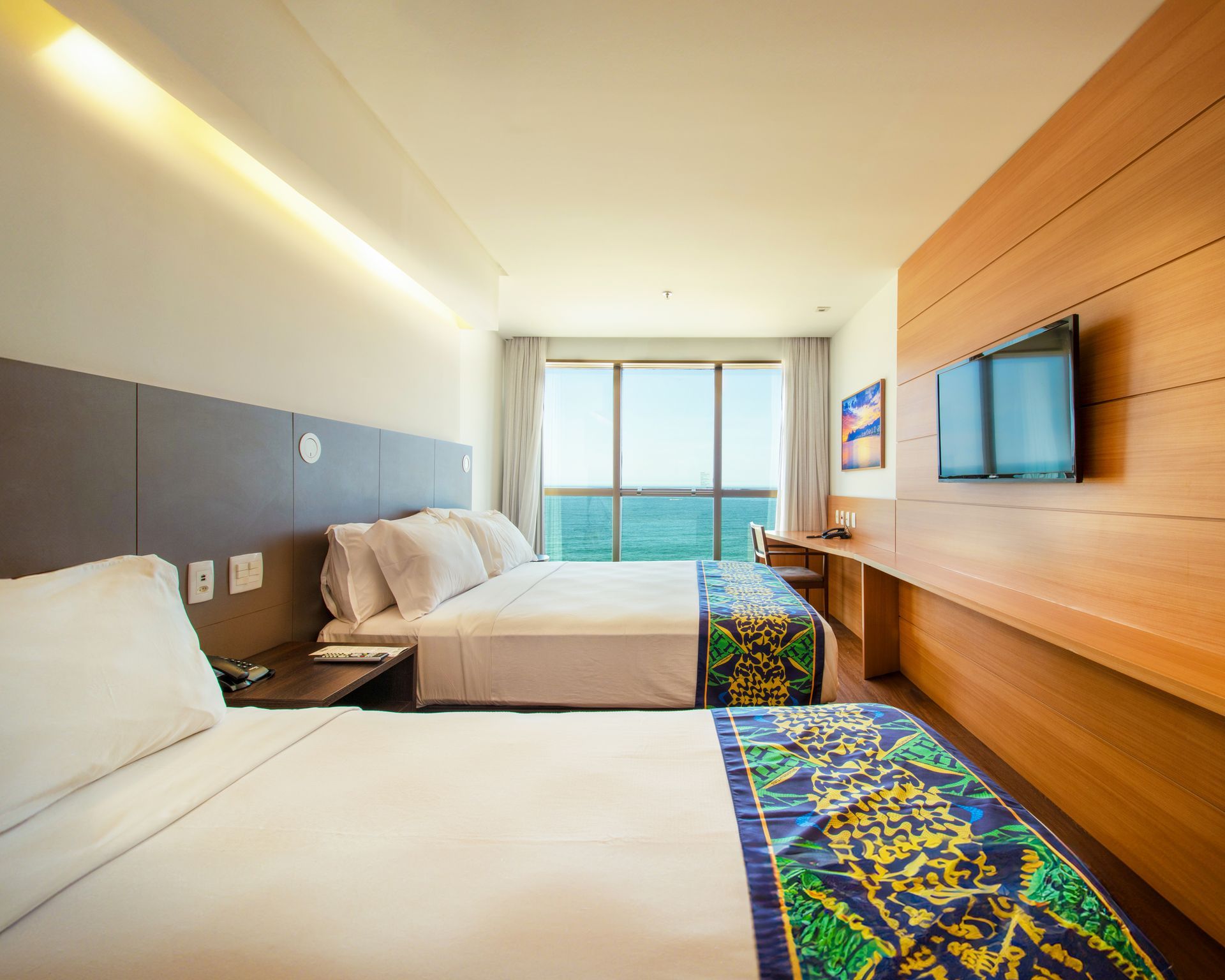 Quarto de hotel moderno com duas camas, vista para o mar, TV de parede e tapetes coloridos.
