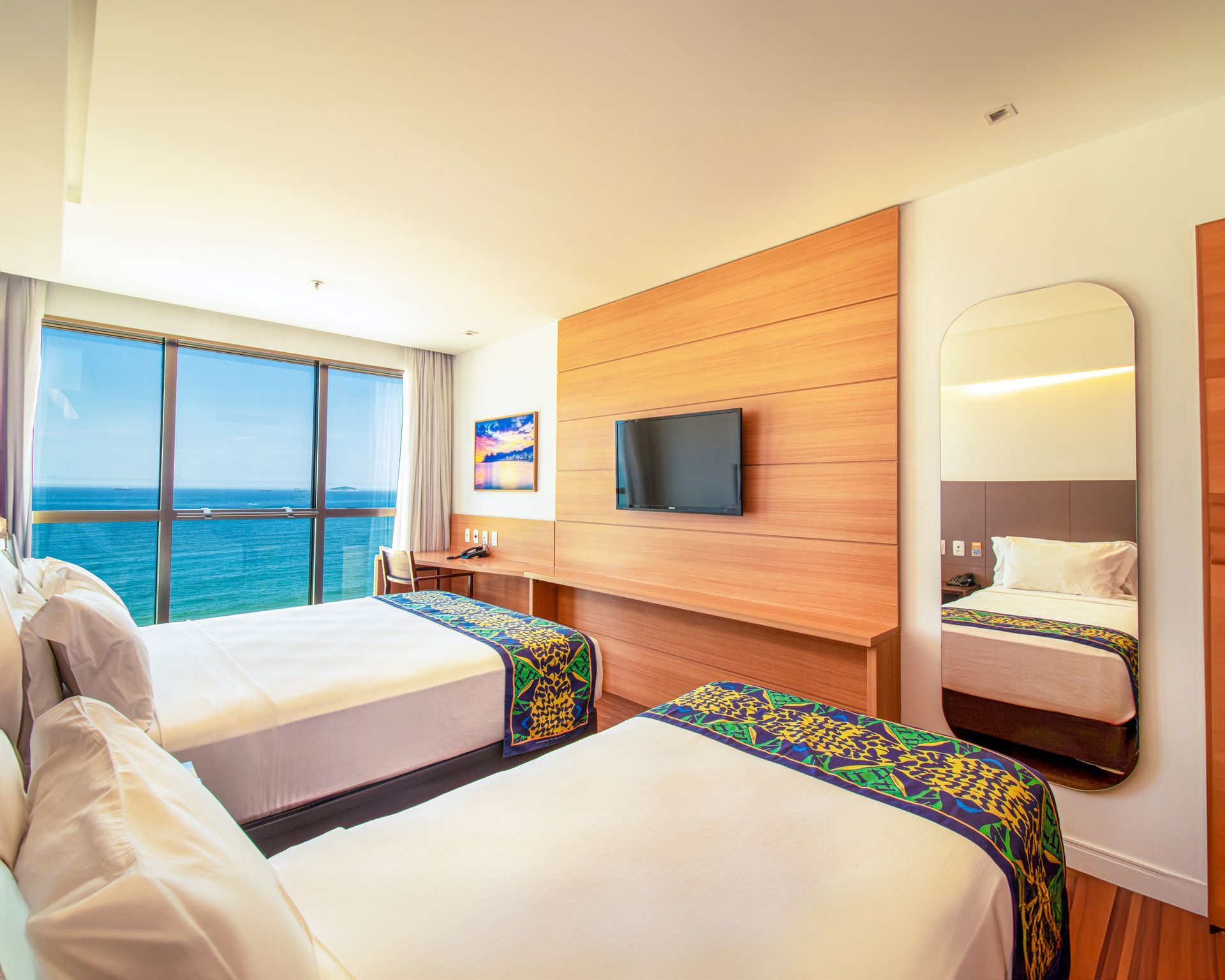 Quarto de hotel luminoso com duas camas, vista para o mar através de grandes janelas, TV de parede e parede com detalhes em madeira.