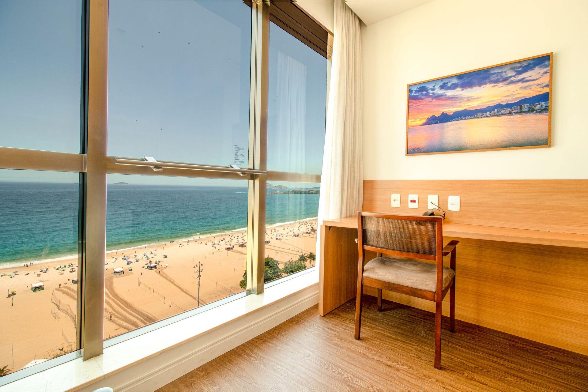 Quarto luminoso com escrivaninha e cadeira, com vista para uma praia ensolarada e para o oceano através de grandes janelas.