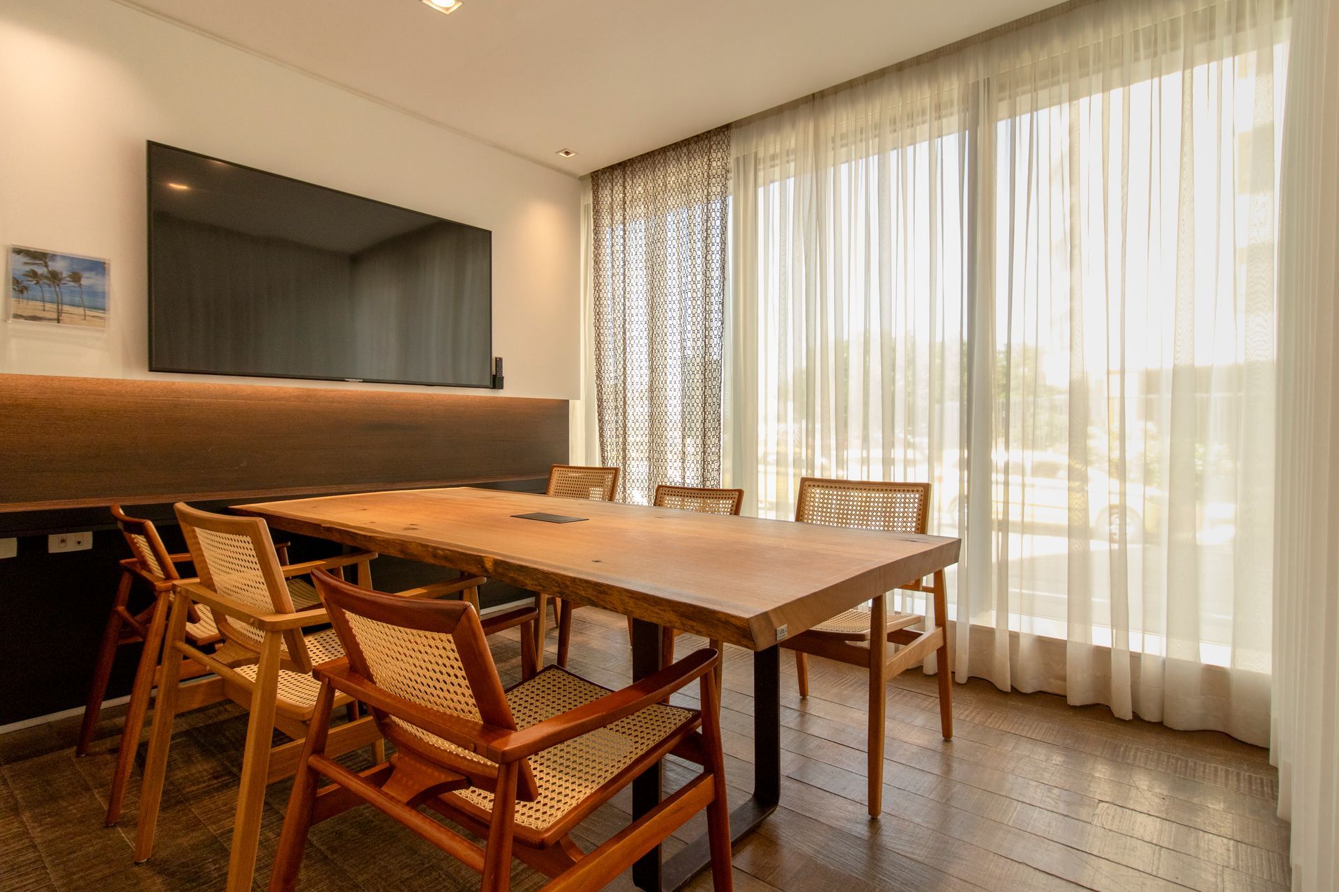 Sala de jantar moderna com mesa e cadeiras de madeira, grande espelho de parede e cortinas translúcidas junto a janelas amplas e iluminadas.