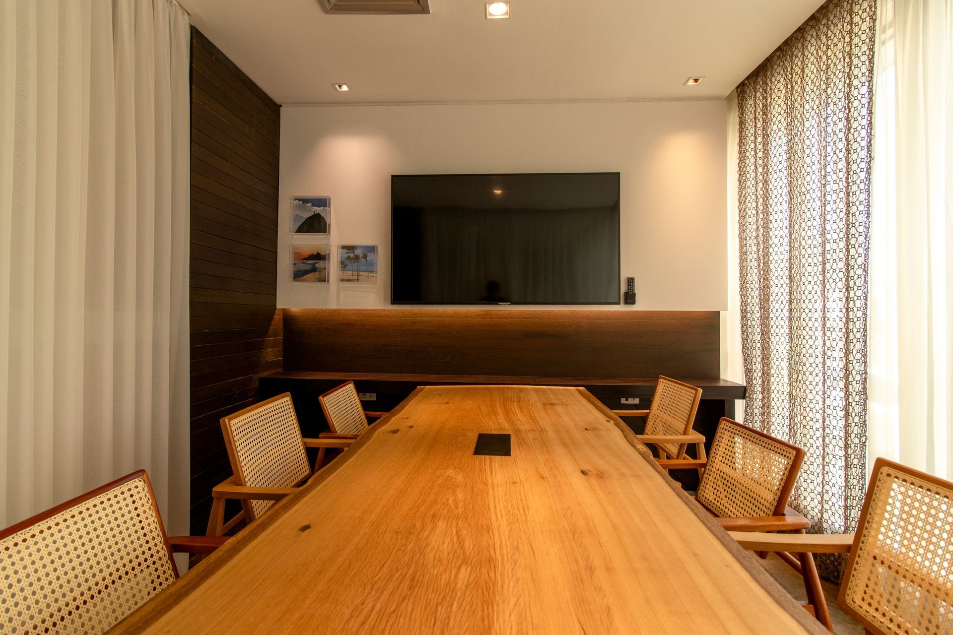 Sala de conferências moderna com mesa comprida de madeira, cadeiras de vime, TV fixada na parede e cortinas estampadas.