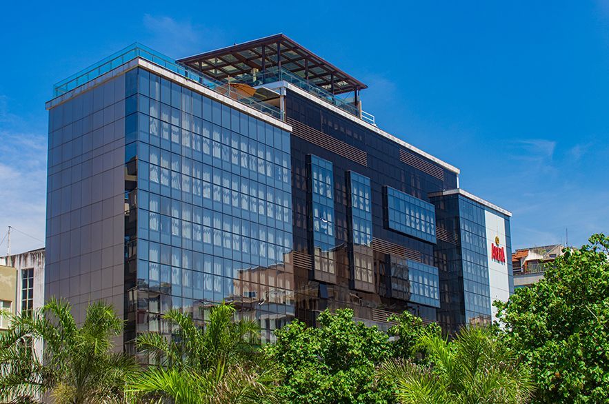 Edifício de escritórios moderno, envidraçado, com estrutura no telhado, emoldurado por árvores verdes sob um céu azul.