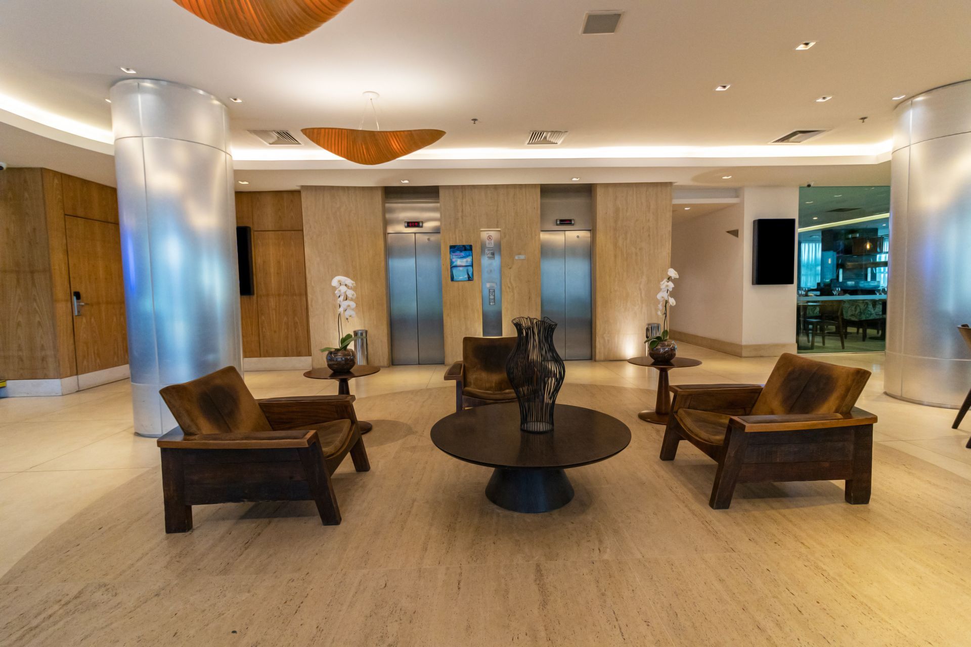 Amplo saguão de hotel com piso bege, detalhes em madeira e poltronas escuras ao redor de uma mesa redonda.