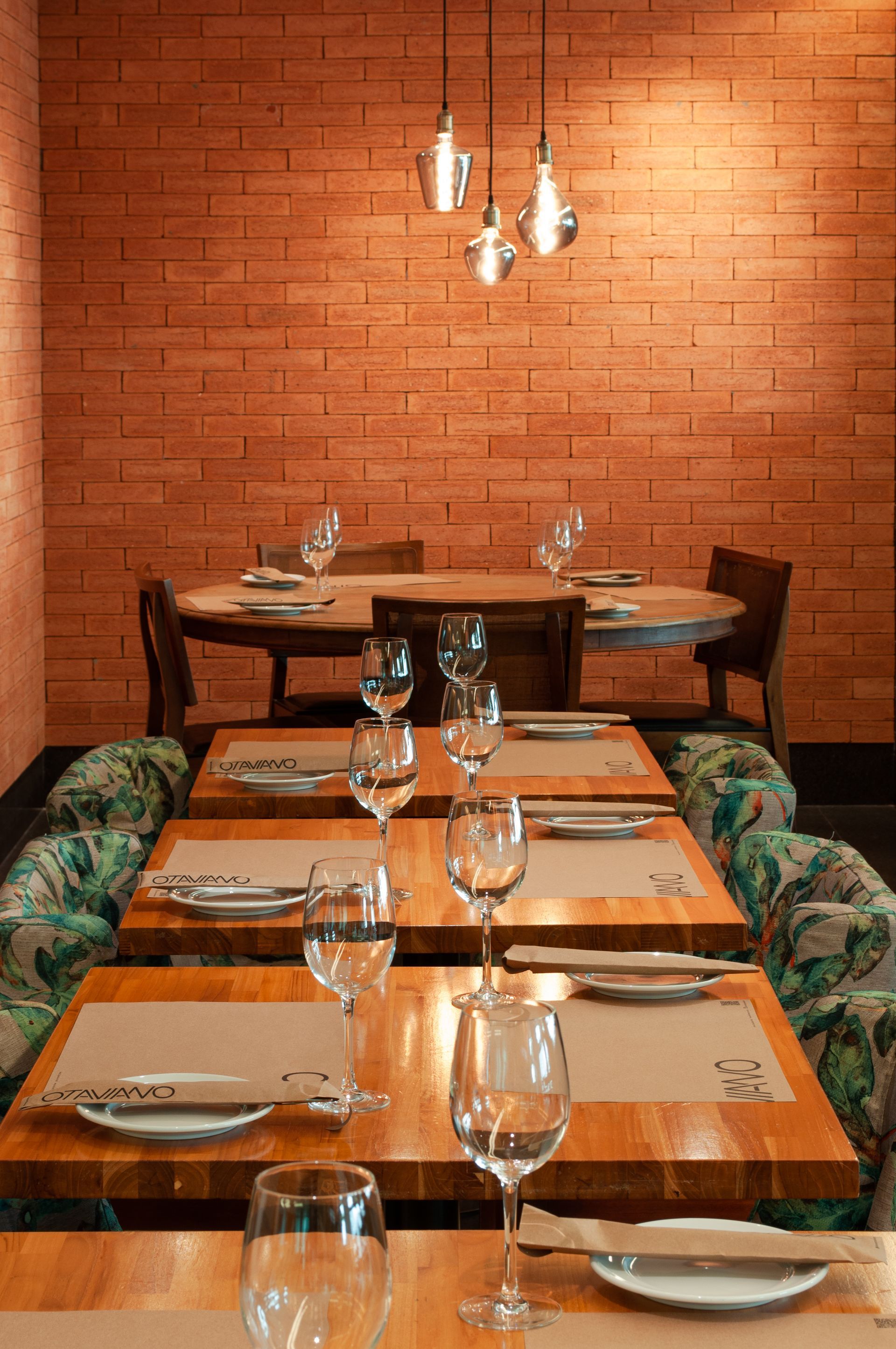 Mesa de restaurante vazia com taças de vinho e jogos americanos encostada em uma parede de tijolos laranja.