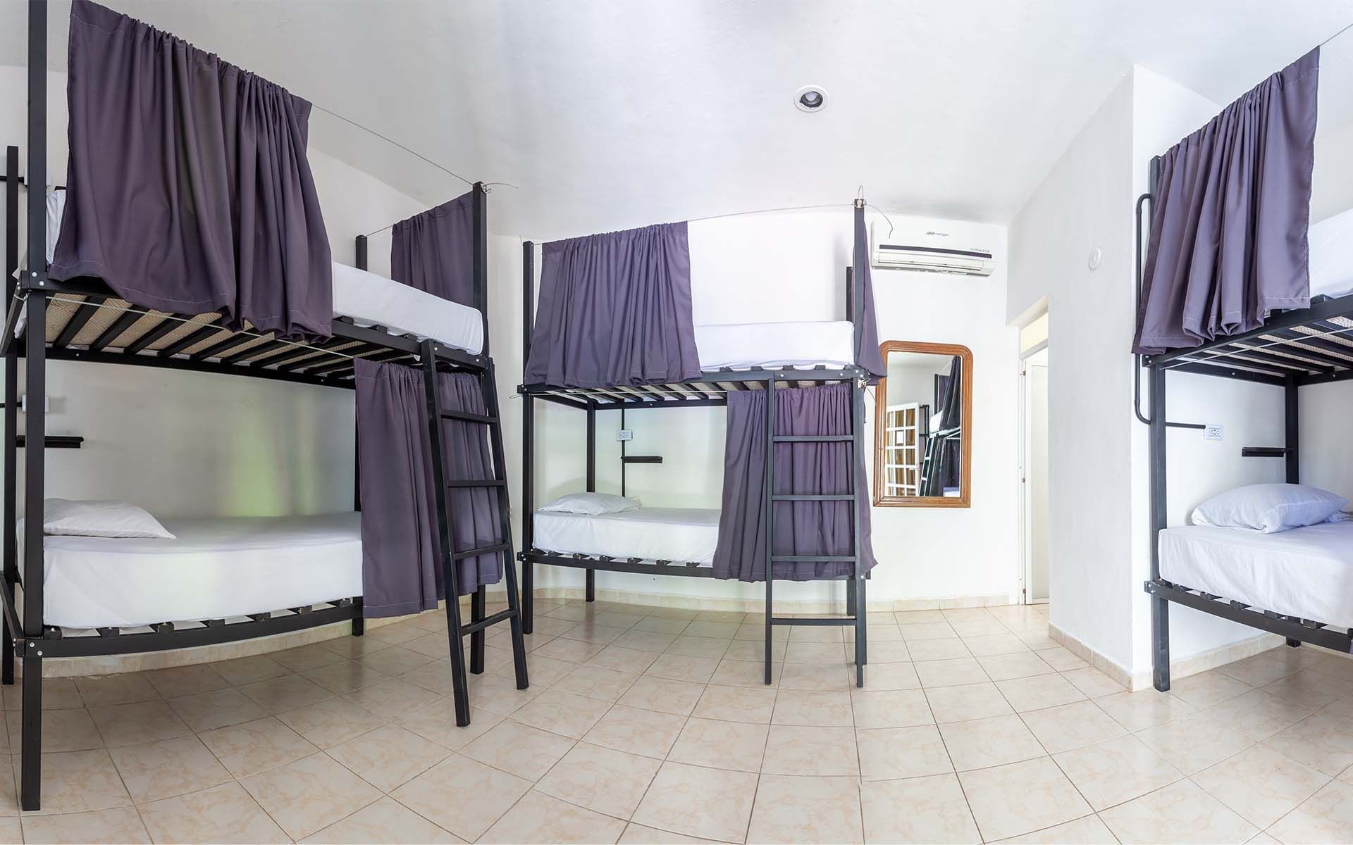 Habitación de albergue con varias literas, paredes blancas y cortinas de color violeta.
