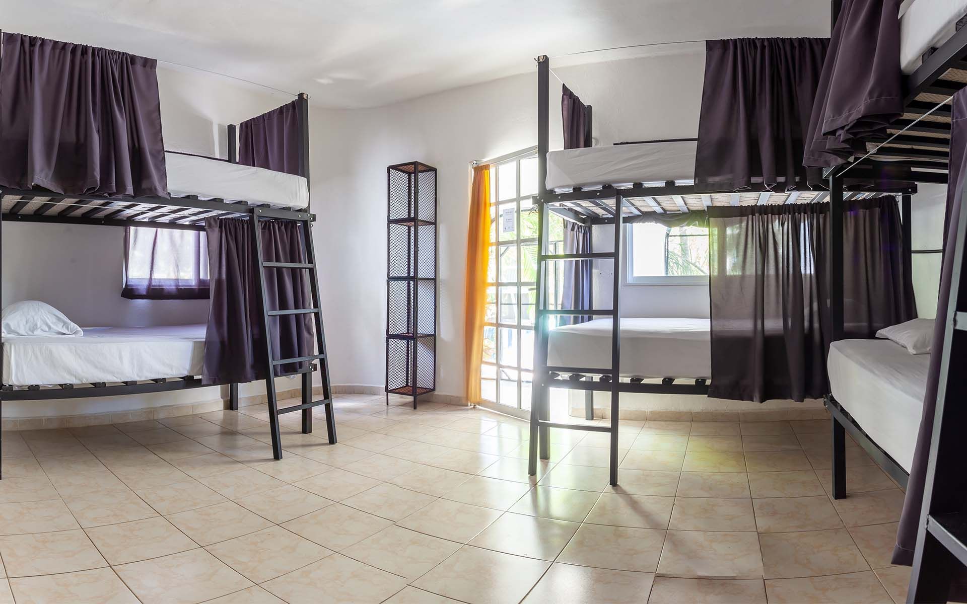 Dormitorio con varias literas, cada una con cortinas. La habitación tiene suelo de baldosas claras y una gran ventana.