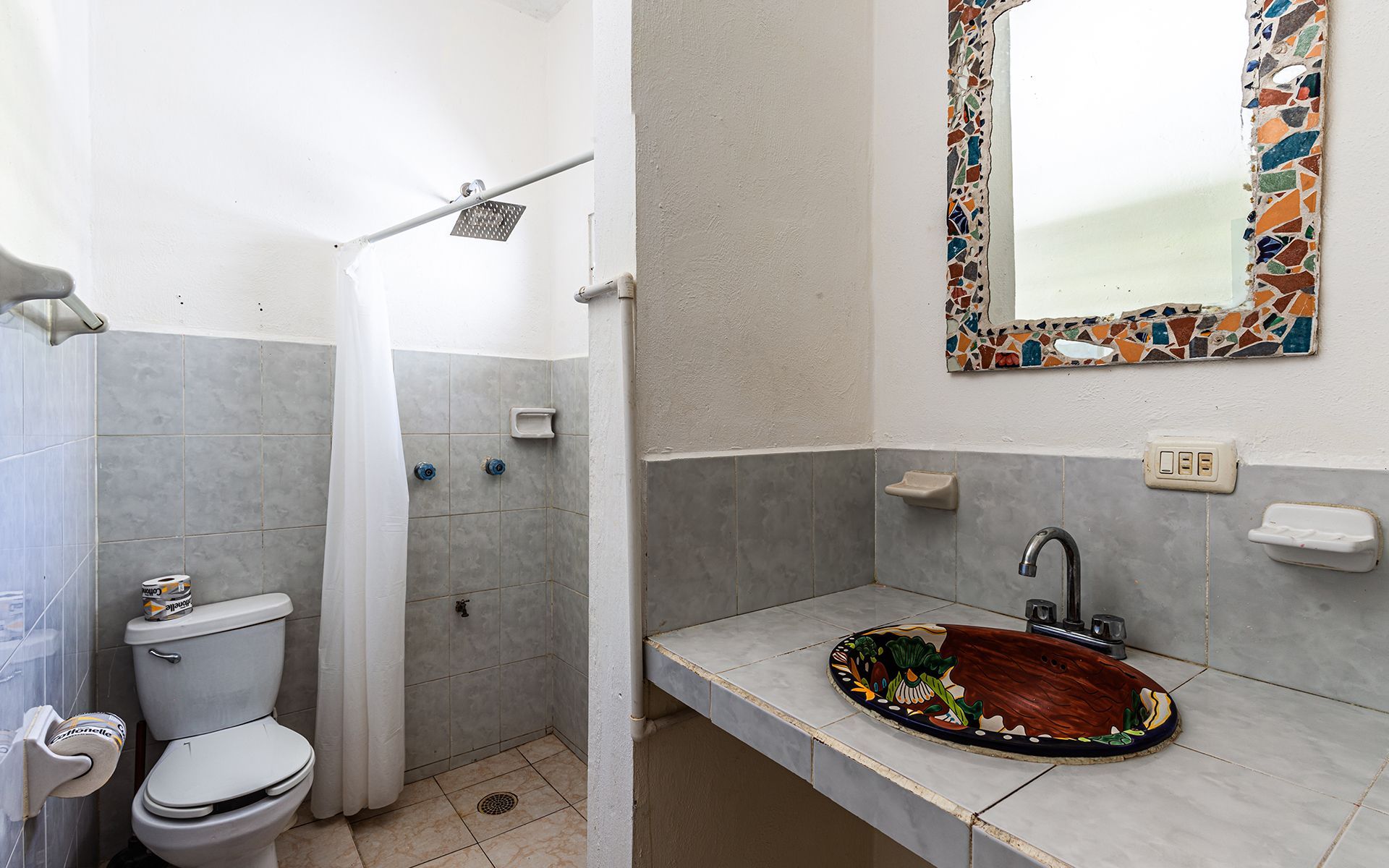 Baño con ducha, inodoro, lavabo y espejo decorativo. Azulejos grises y paredes blancas.
