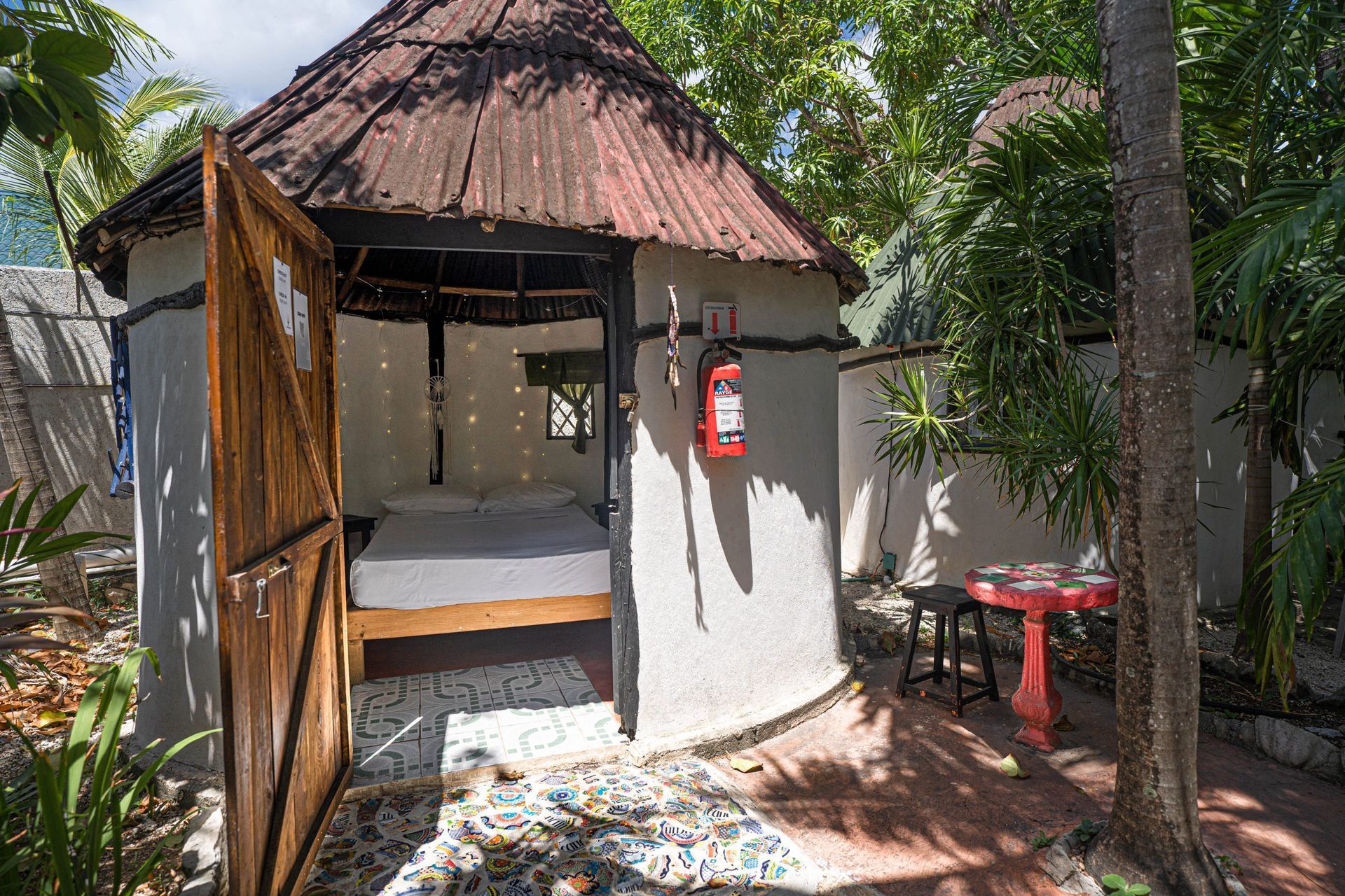 Una pequeña cabaña redonda con una puerta de madera abierta, una cama adentro y una mesa roja y un taburete afuera.