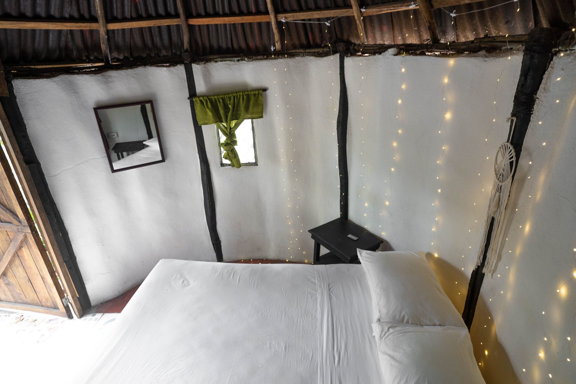 Interior de una habitación rústica con cama, ventanas pequeñas y luces de colores. Paredes blancas, vigas de madera.