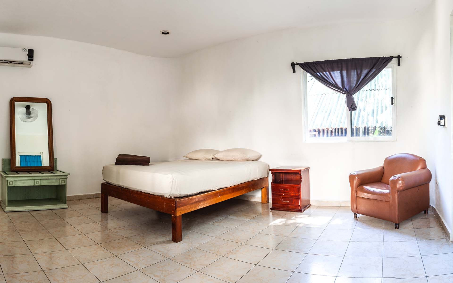 Dormitorio con cama, mesita de noche, sillón, cómoda y ventana con cortina oscura.