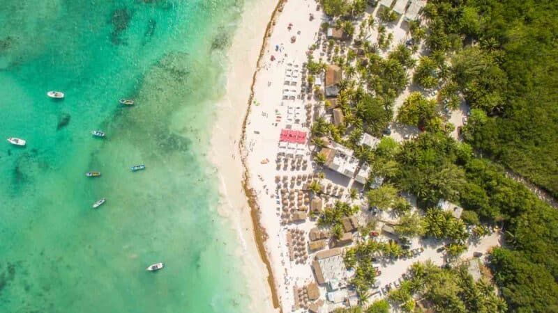 Vista aérea de una playa de arena blanca con agua turquesa, barcos y un exuberante bosque verde.