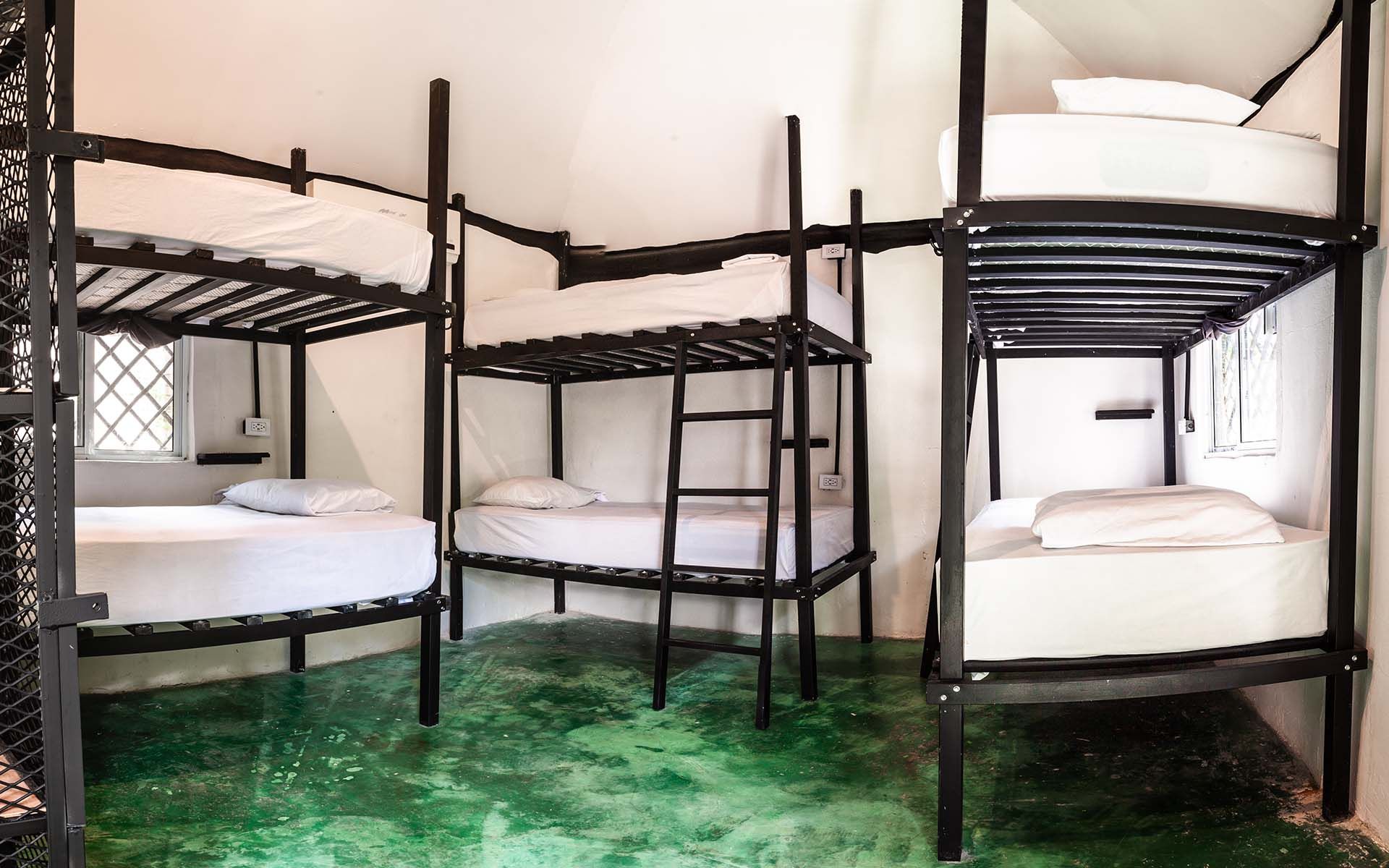 Habitación de albergue con tres literas metálicas. Ropa de cama blanca, suelo verde y ventanas pequeñas.