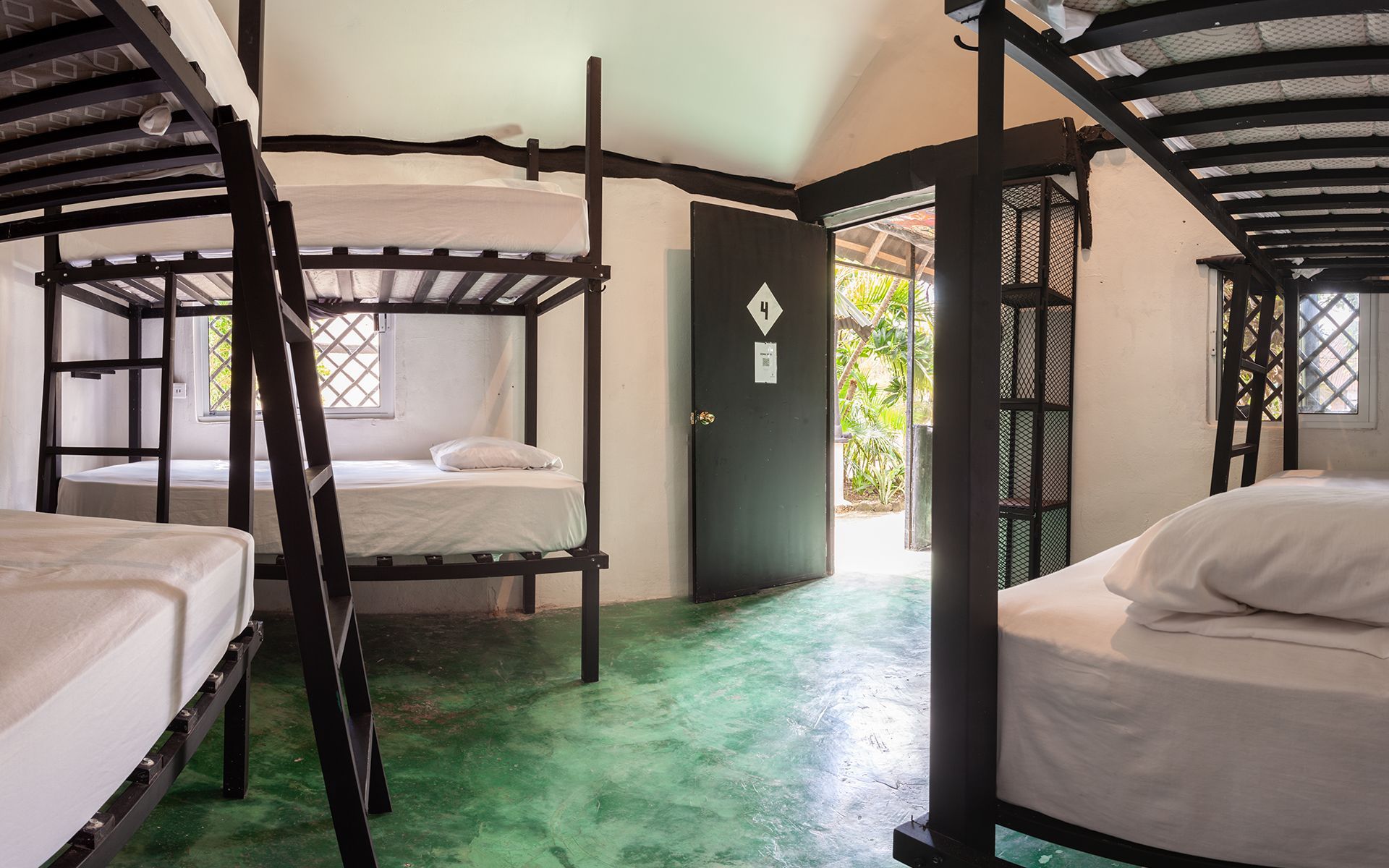 Habitación compartida en albergue con varias literas, puerta al exterior, suelo verde, paredes blancas y luz natural .