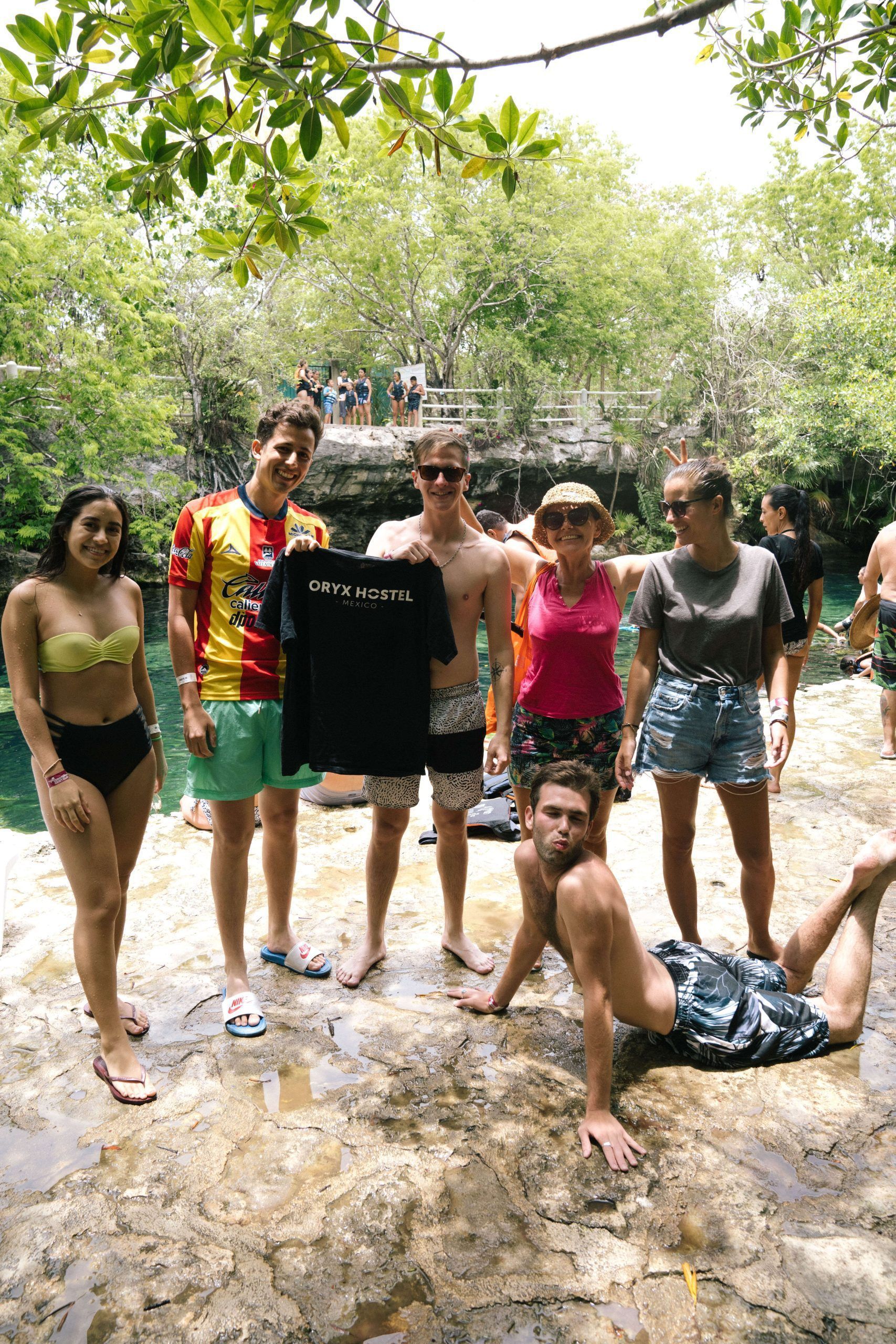 Grupo de personas cerca de un cuerpo de agua con una camiseta negra. La luz del sol se filtra entre los árboles.