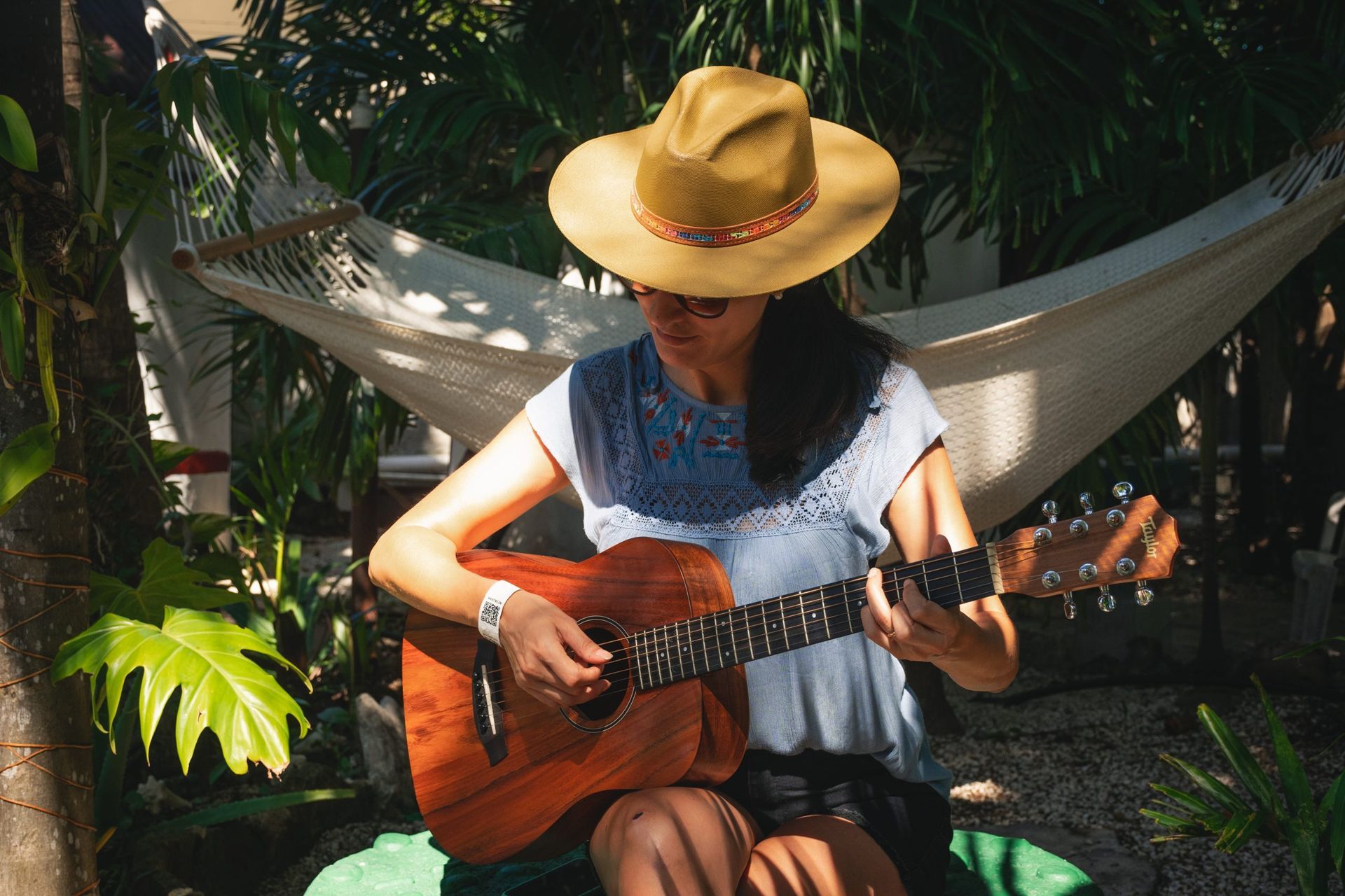 Una mujer con sombrero toca una guitarra acústica marrón al aire libre, con una hamaca de fondo.