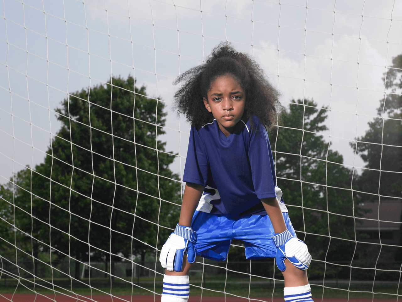 Young soccer goalkeeper in blue gear crouches behind a goal net on a field