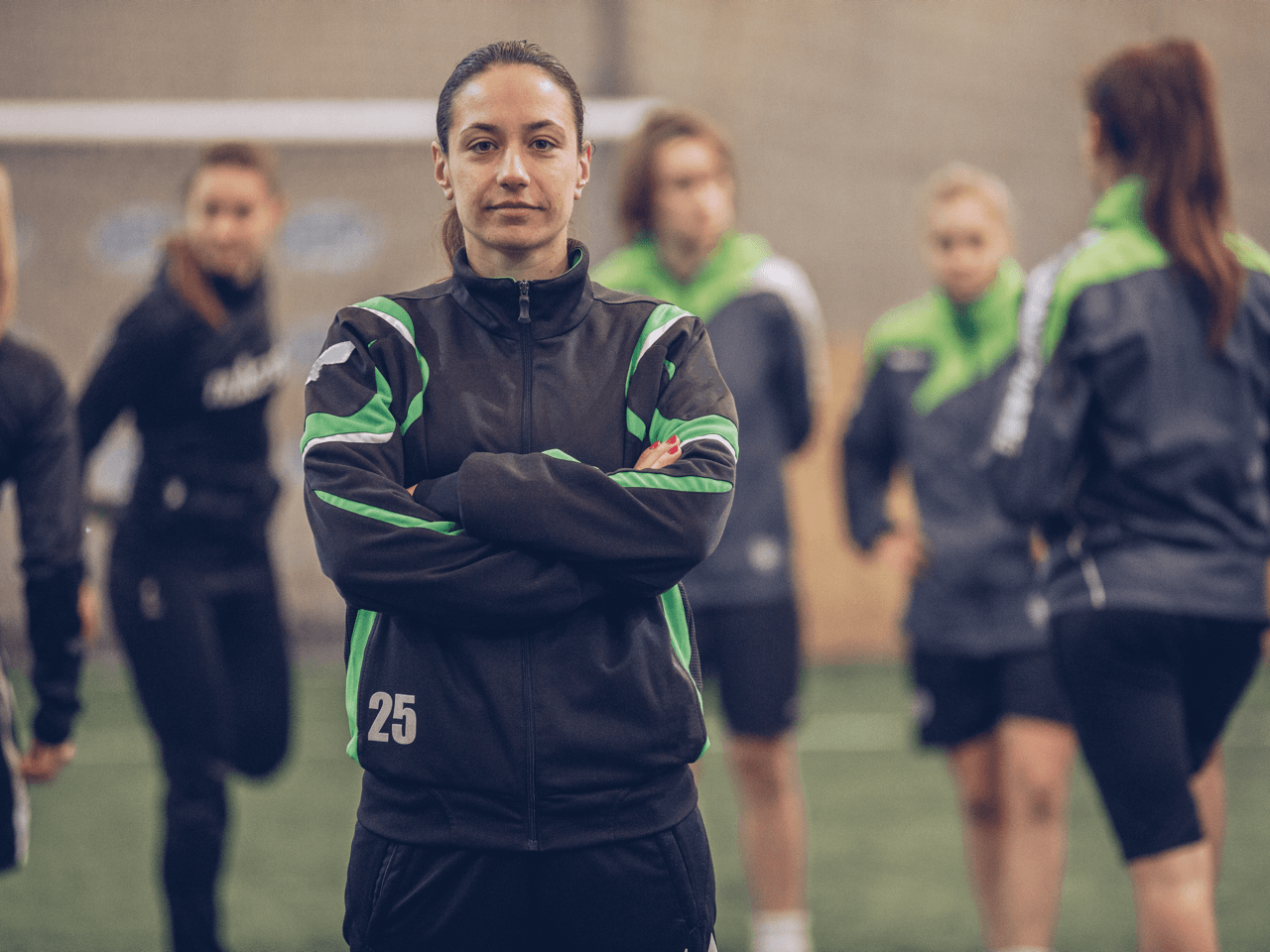 Soccer coach standing with arms crossed on a field, players blurred in the background