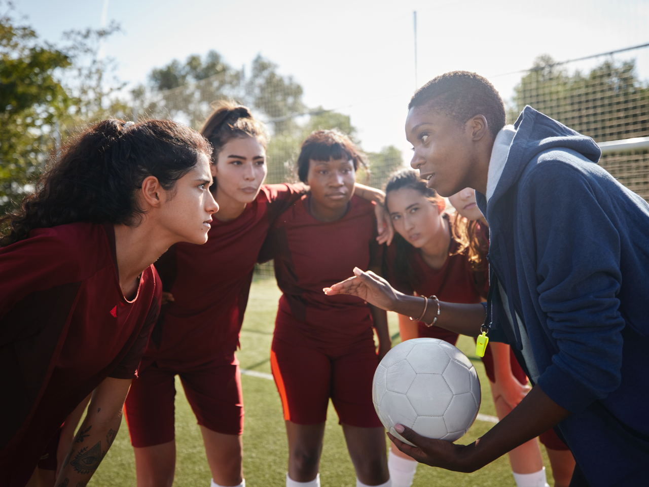 Soccer team huddles on field with coach holding a ball, discussing strategy before practice