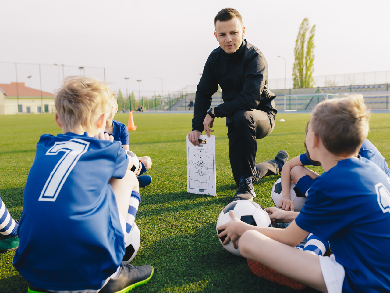 Youth soccer coach kneels with players on a grassy field, discussing tactics around a clipboard.