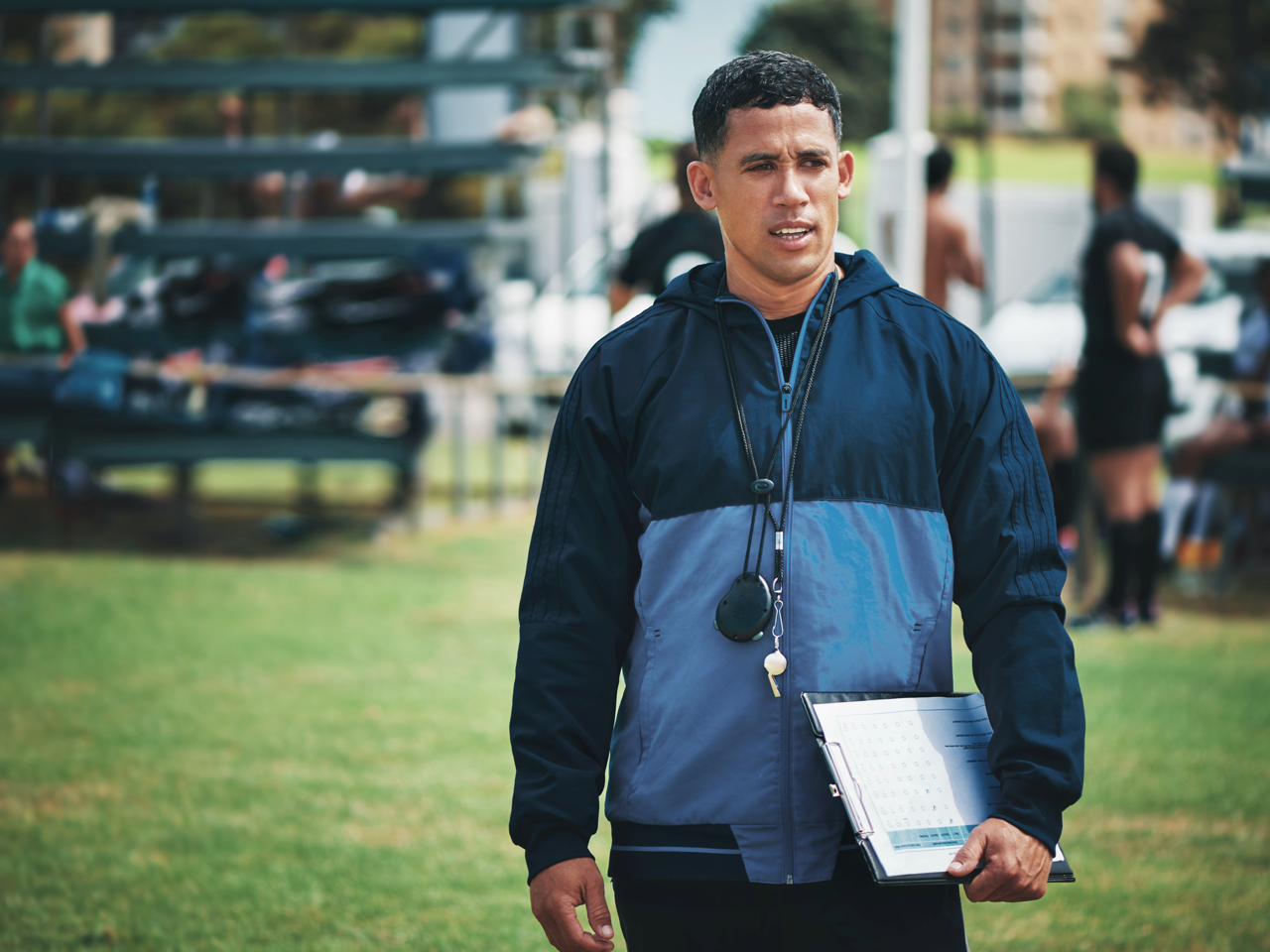 Man standing on a sports field holding papers, wearing a navy and blue jacket with a whistle around his neck.