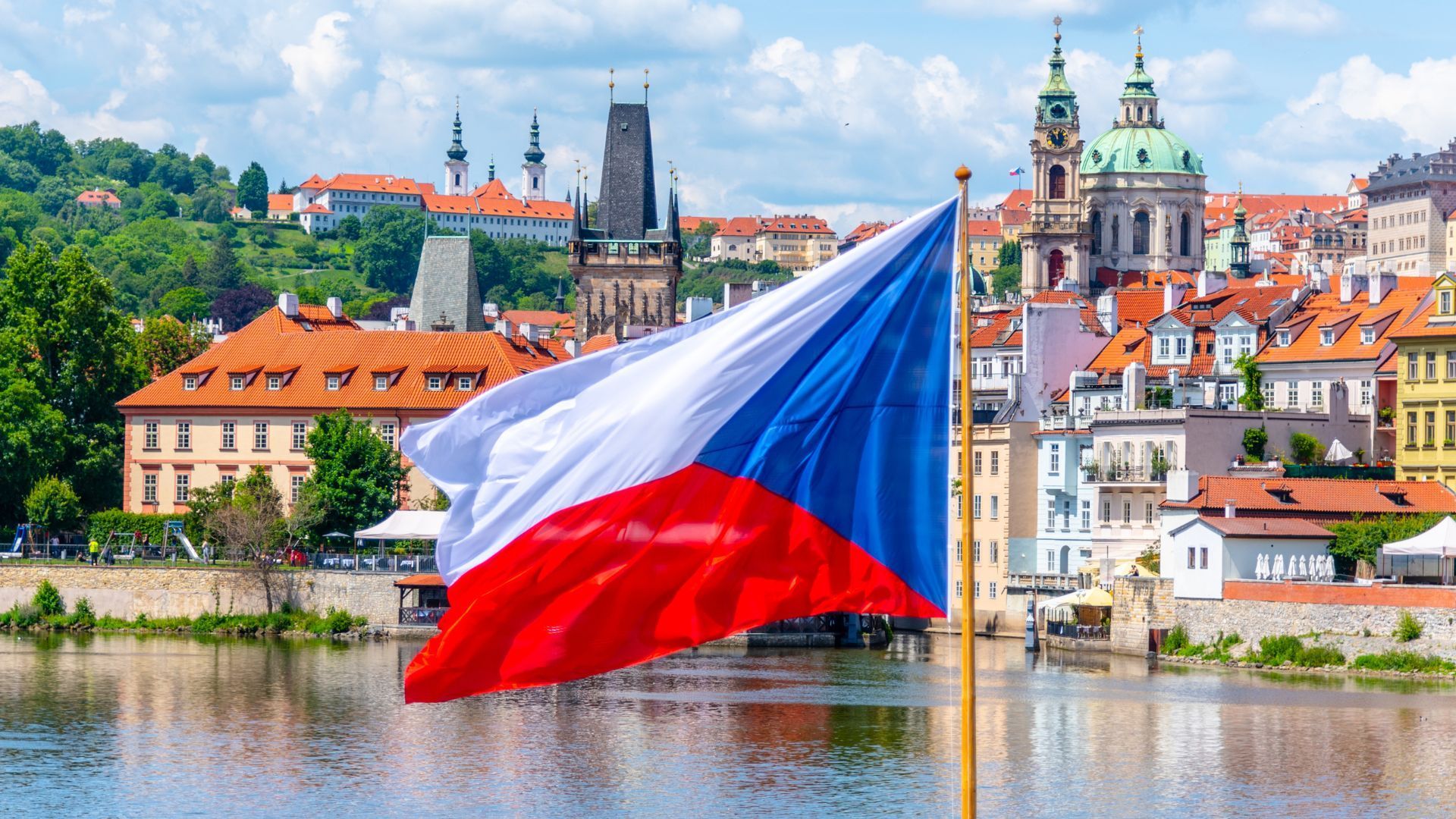 Czech flag waving over Prague cityscape with colorful buildings and river.
