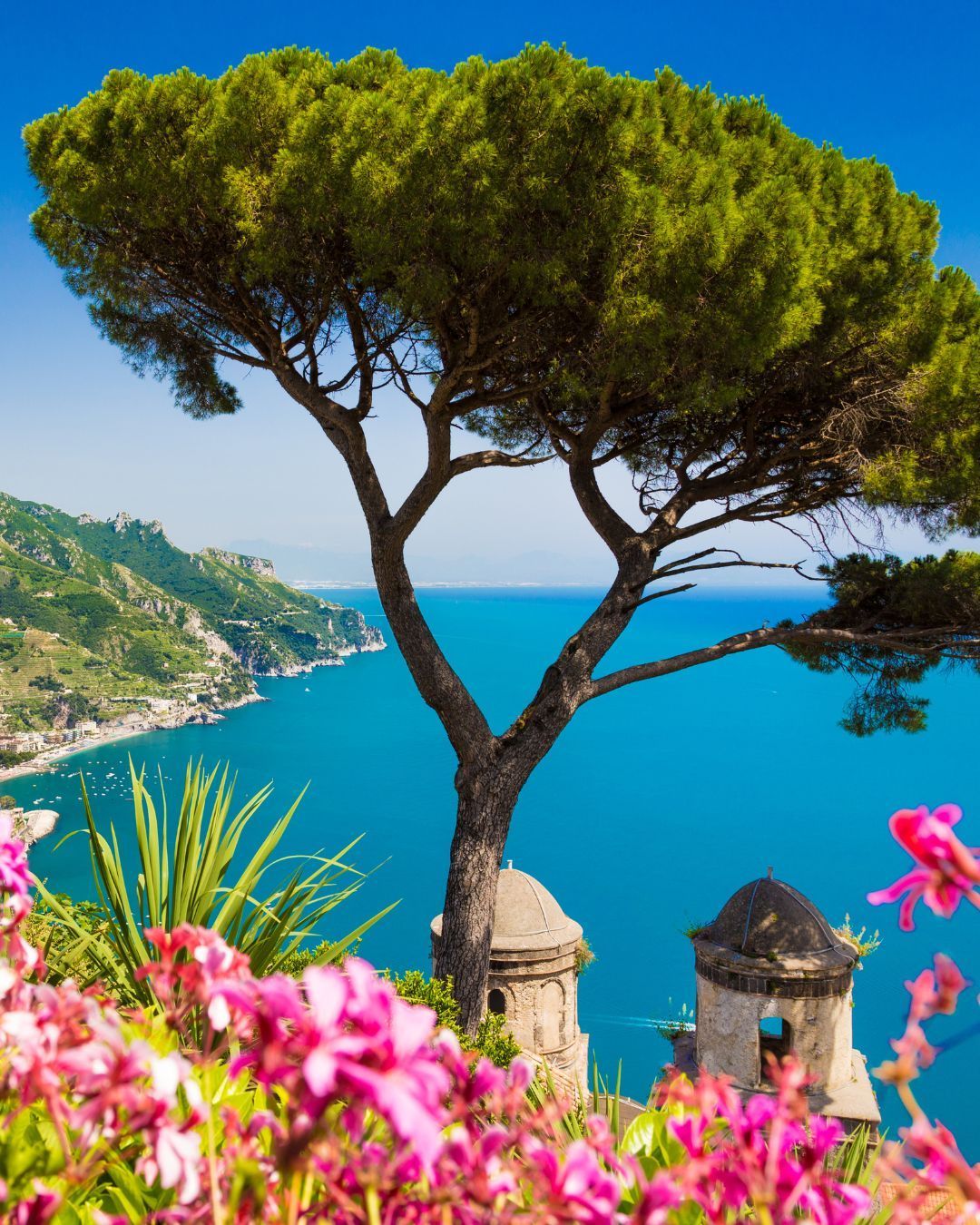 White terrace overlooking a bright blue sea with distant islands and potted flowers in the foreground
