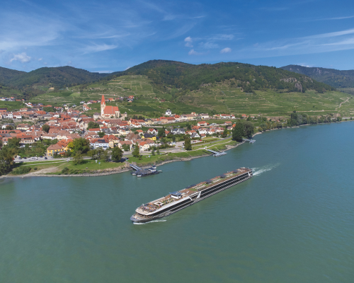 Travelmarvel Capella river cruise ship passes a riverside town with red-roofed buildings and green hills under a blue sky.